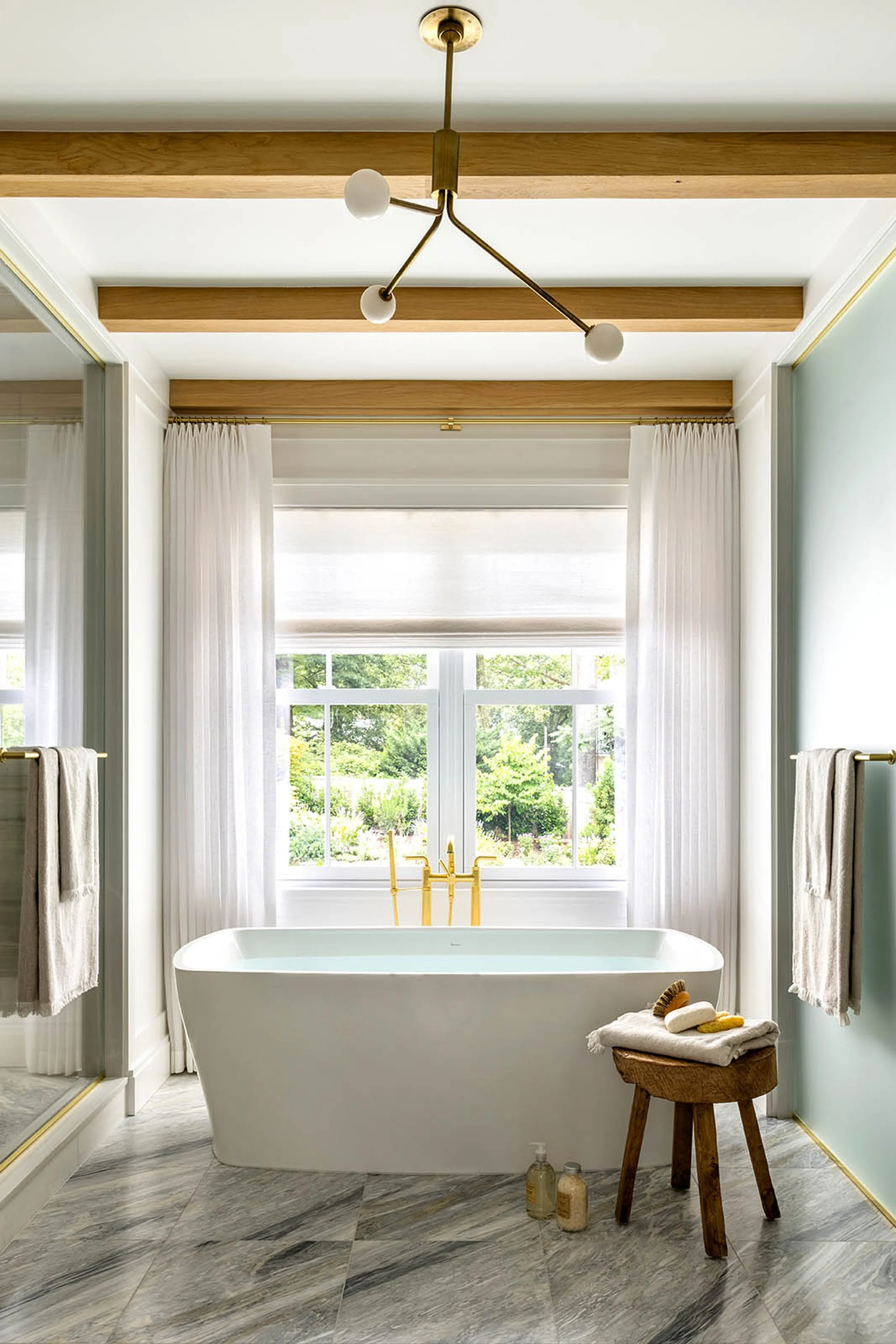 A modern bathroom with a freestanding bathtub positioned in front of a large window with white curtains. The bathroom has wooden beams on the ceiling, marble flooring, and a gold chandelier. There is a small stool next to the bathtub with folded towe