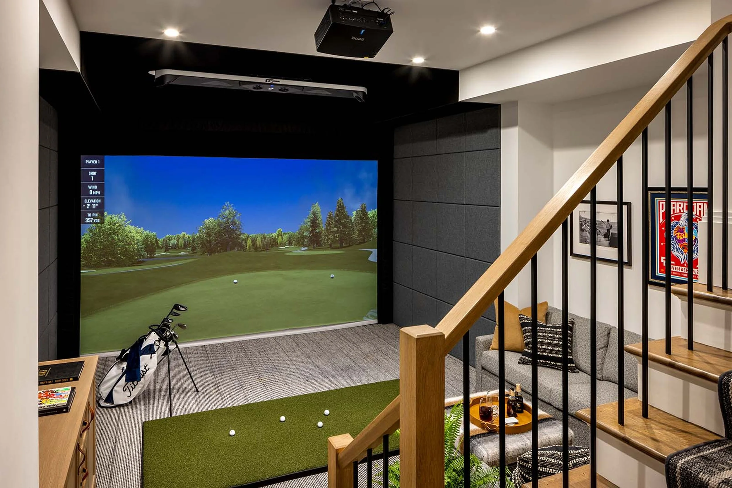 Indoor golf simulation room with a large screen displaying a golf course, golf clubs, and a golf bag on the floor, with framed pictures on the wall and a small table with drinks and snacks nearby.