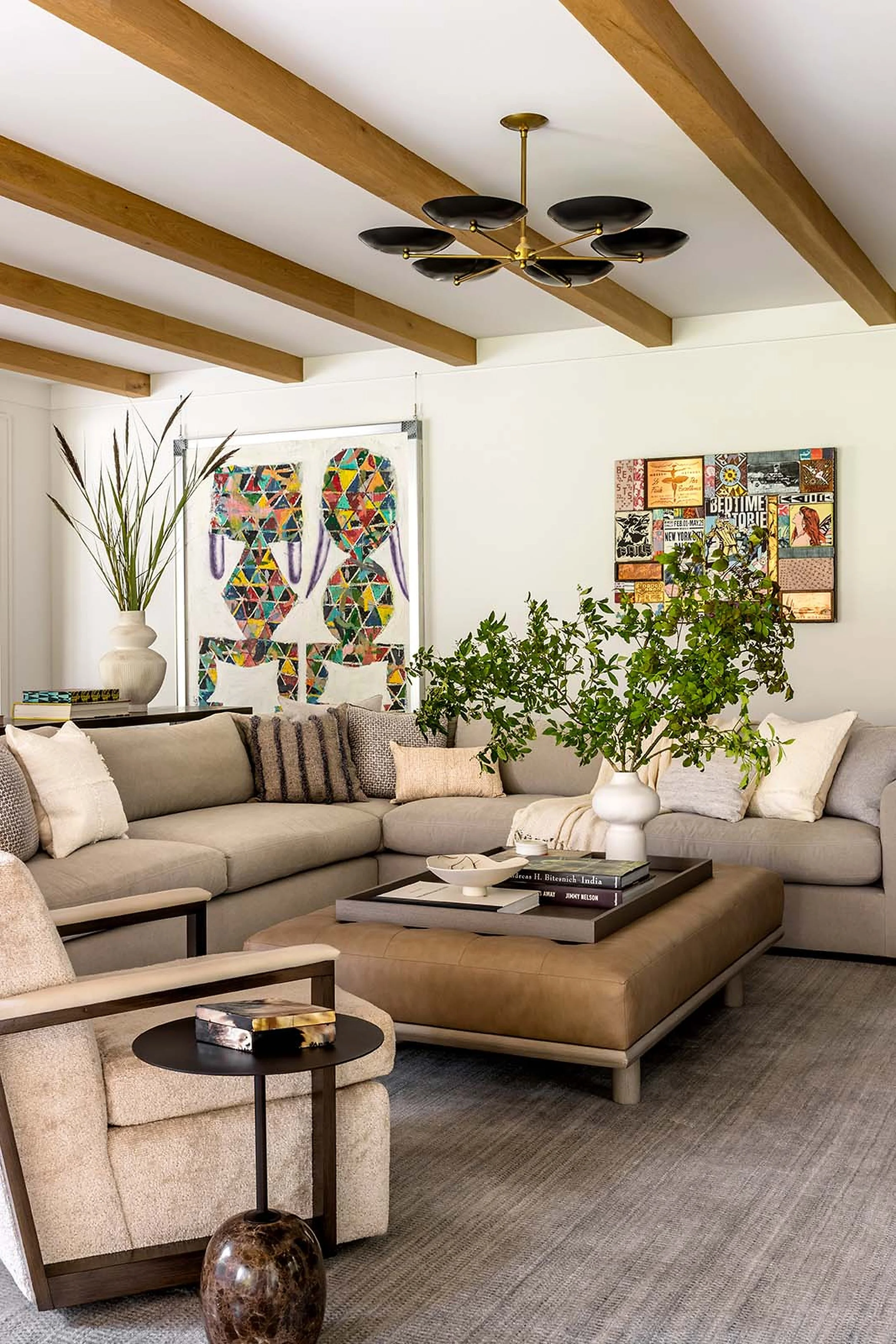 Living room with beige sectional sofa, wooden coffee table, black side table, linen and textured cushions, potted green plant, abstract artwork on the wall, and ceiling with wooden beams and modern black and gold chandelier.