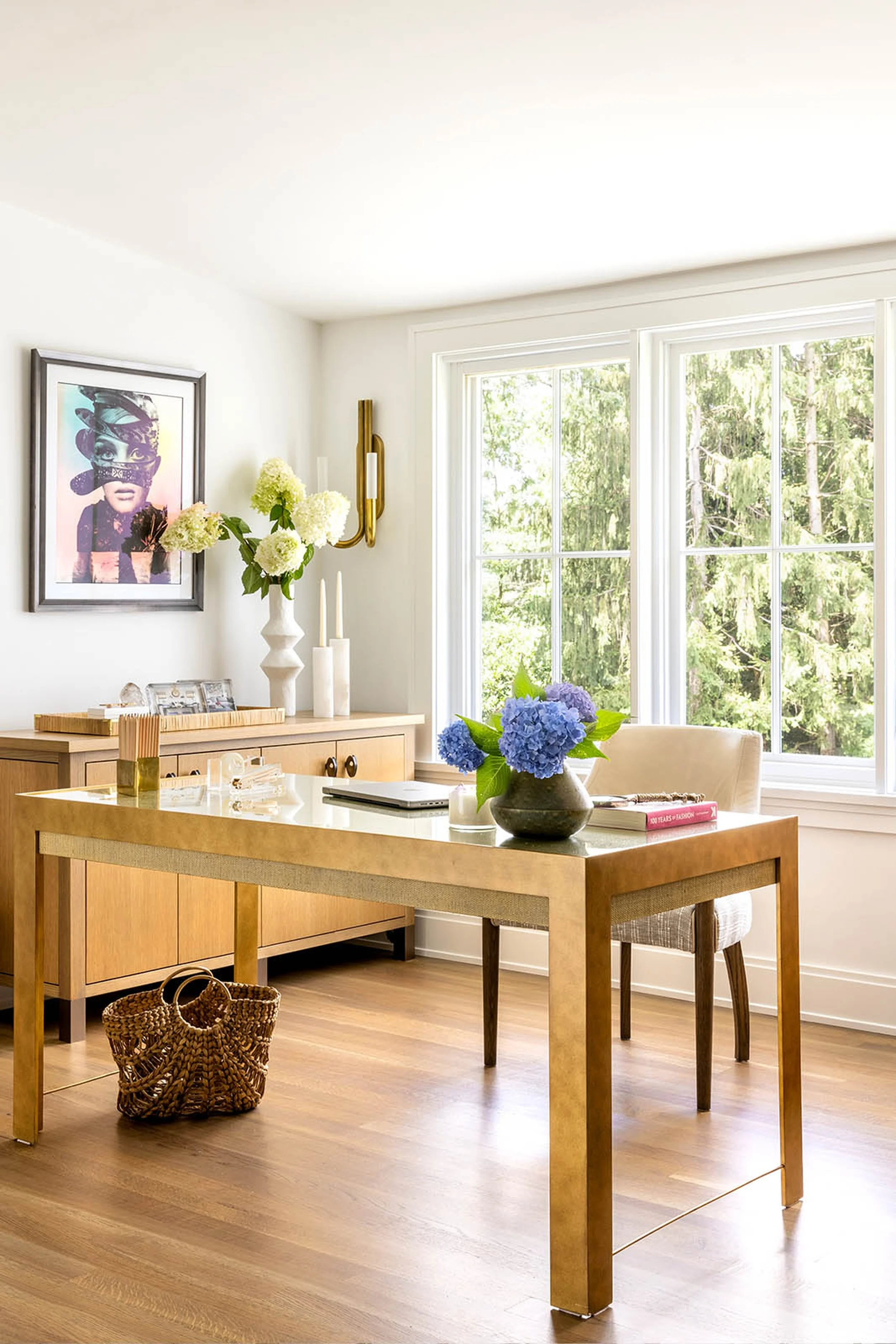 The image shows a cozy, well-lit kitchen or dining area with a wooden table and matching chairs. On the table, there is a dark vase with bright blue hydrangea flowers, a closed laptop, and a pink book. Behind the table, a console or sideboard display