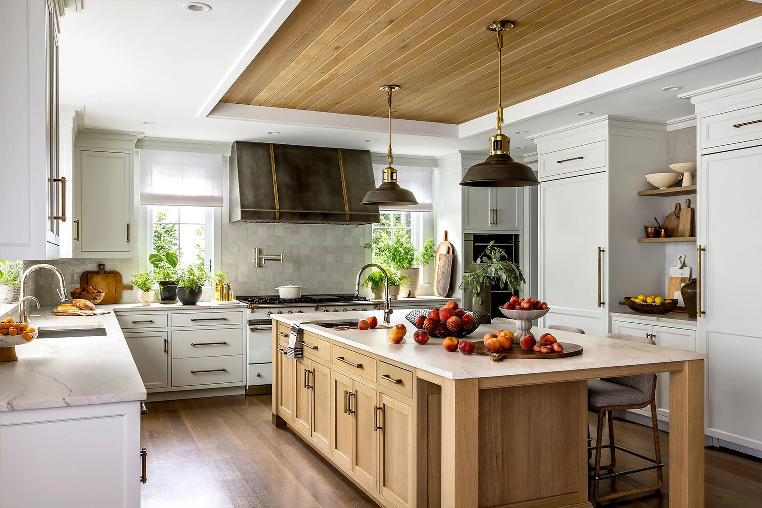 A modern kitchen with white cabinets, a wooden kitchen island, and a wooden ceiling. The kitchen is decorated with fresh peaches in bowls and on the counter, green plants, and kitchen utensils.