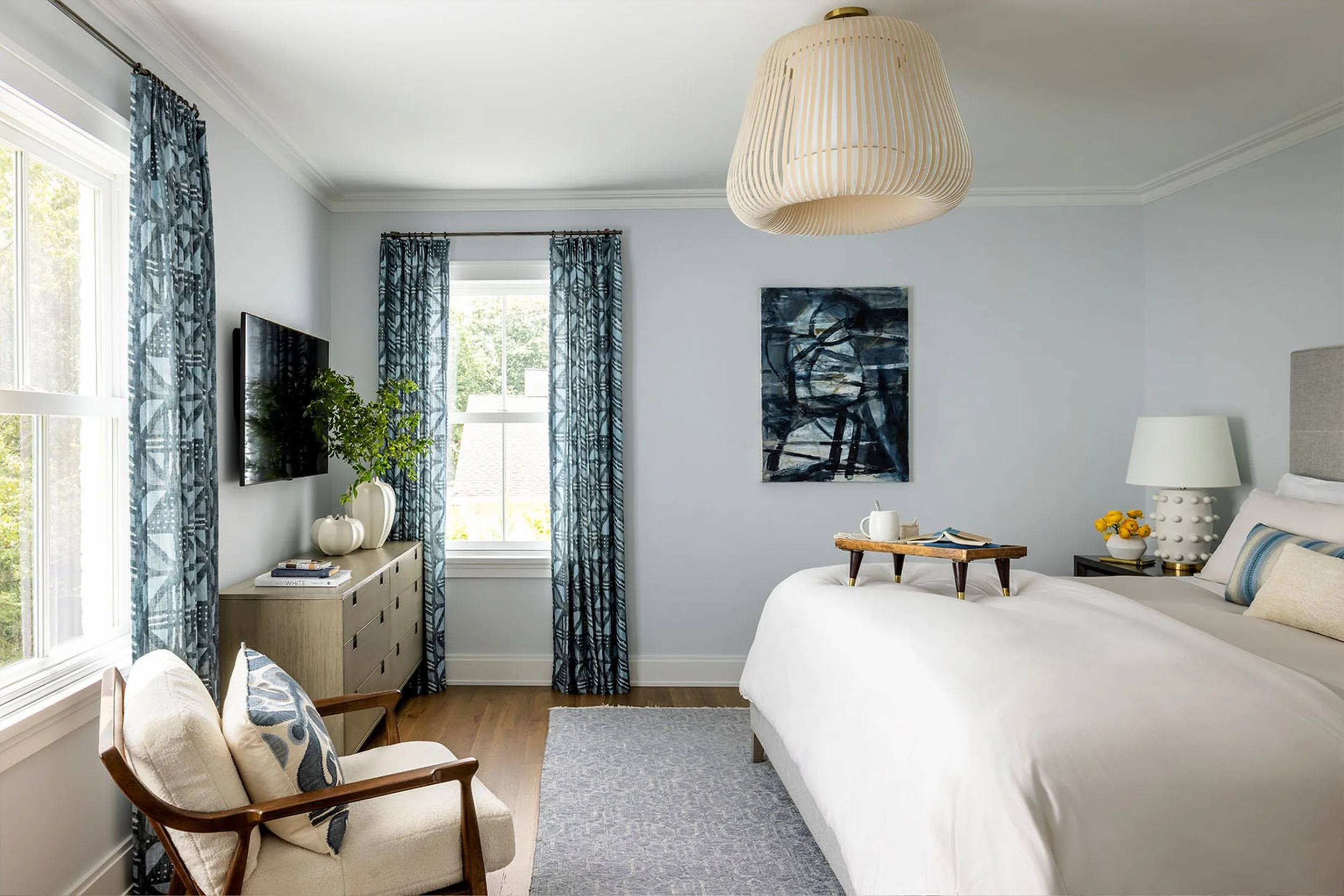 A cozy bedroom with a white bed, blue patterned curtains, a wooden chair with beige cushion and blue pillow, a gray dresser with books and a plant, a black nightstand with a white lamp and yellow flowers, abstract art on the wall, a large paper penda