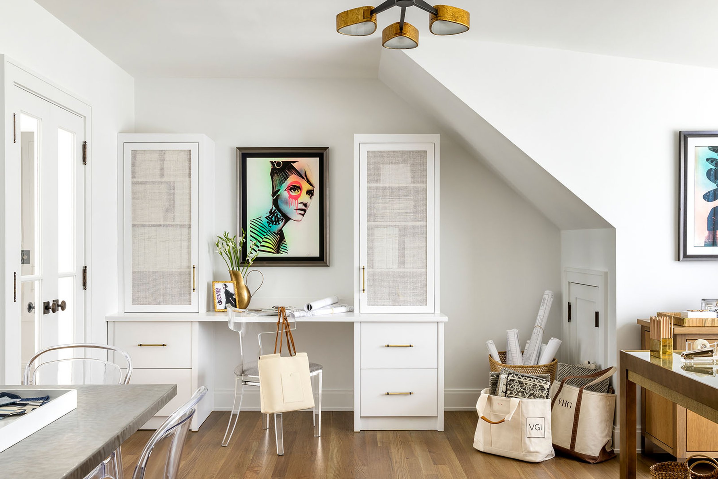 A white room with a sloped ceiling, decorated with artwork including a framed colorful portrait of a woman. There are shelves, a table, chairs, and various miscellaneous items, including bags and rolled-up papers.