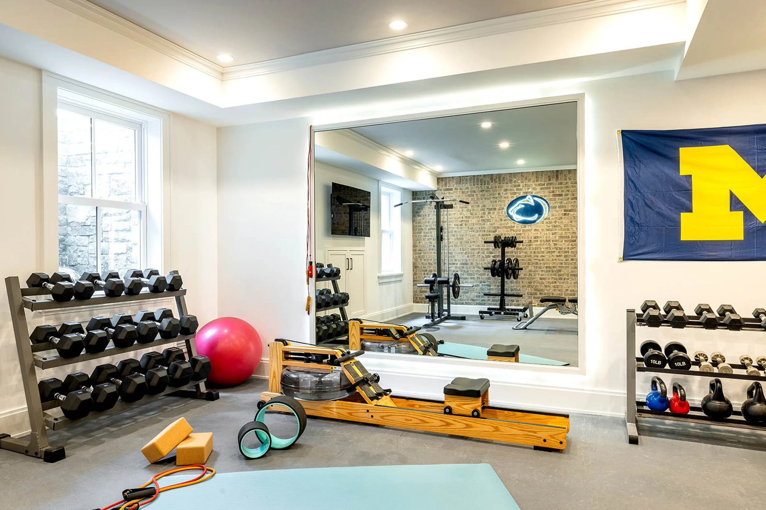 Home gym with dumbbells on racks, kettlebells, exercise mats, foam rollers, resistance bands, a punching bag, and a wall-mounted television.