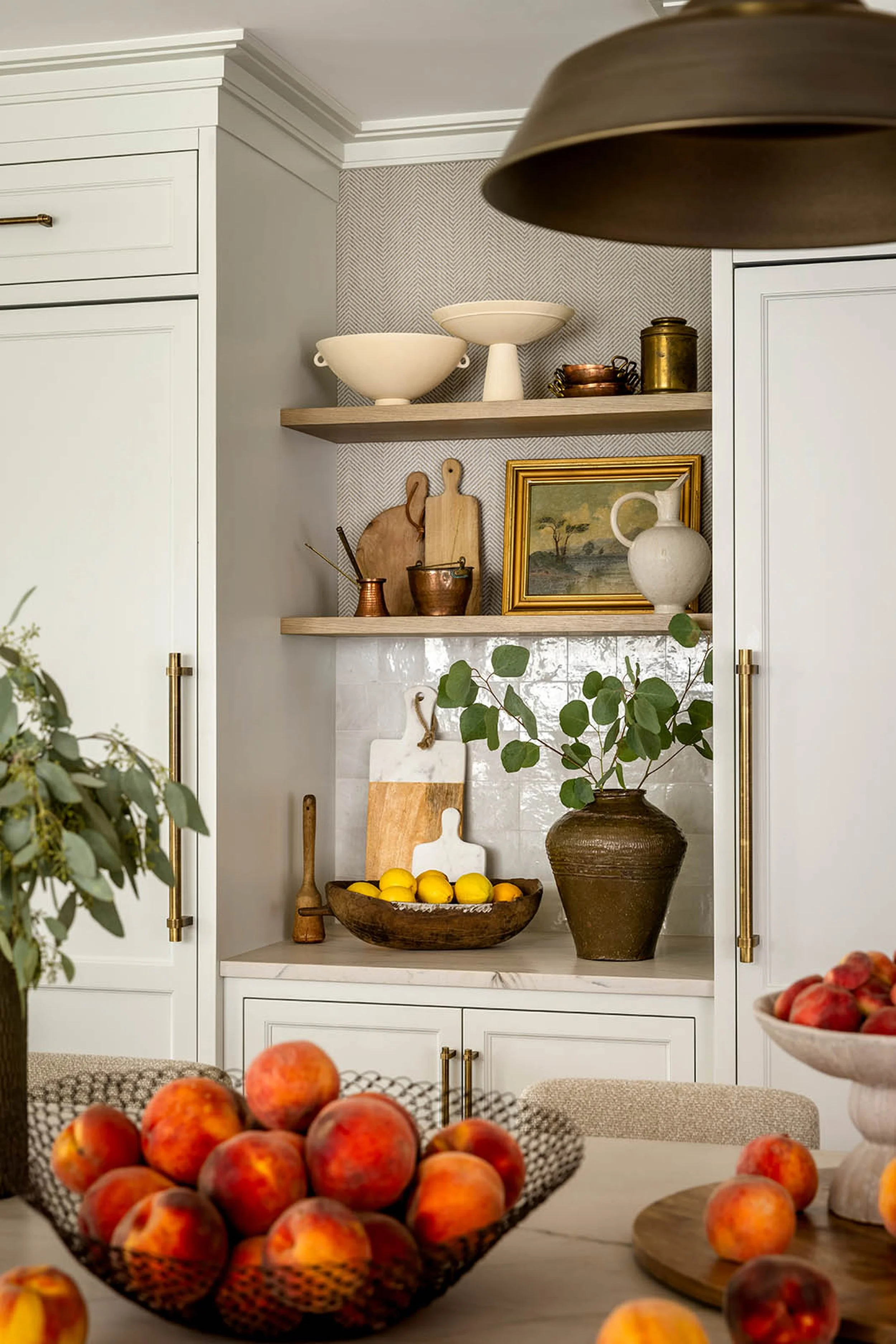 A kitchen cabinet with two open shelves displaying ceramic bowls, copper pots, and a framed landscape picture. A large brown vase with green leaves stands nearby. Apples in a bowl and on the counter are visible in the foreground.