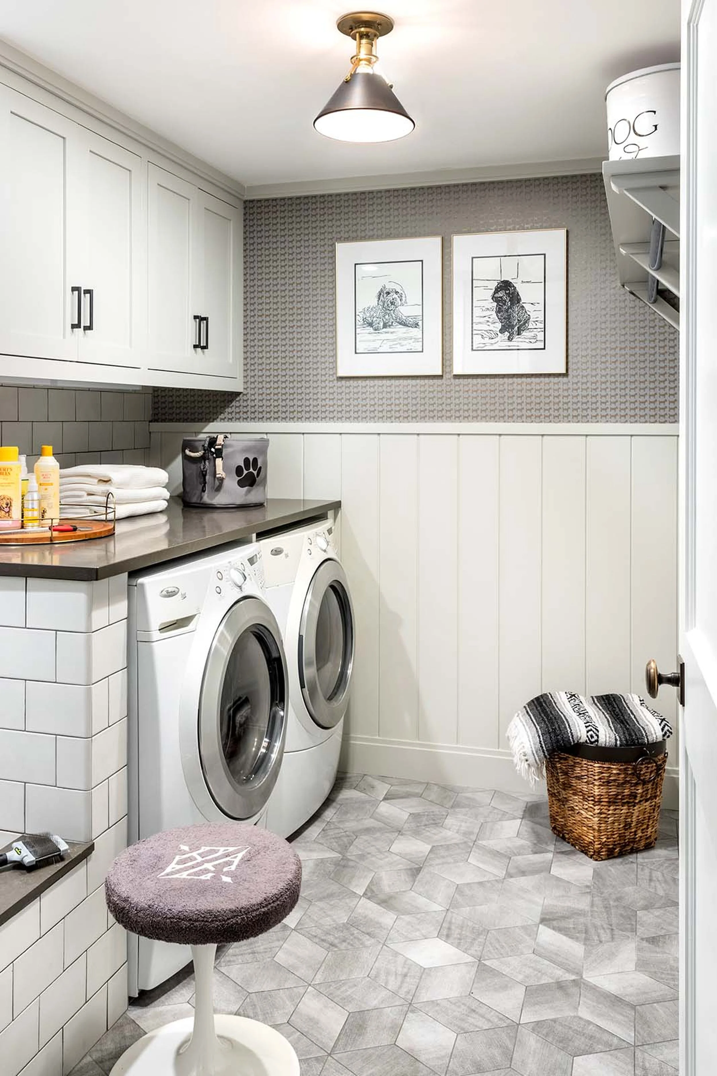 A laundry room with white cabinets, a washer and dryer, framed dog artwork on the wall, a small upholstered stool, a woven laundry basket, and laundry supplies on a counter.