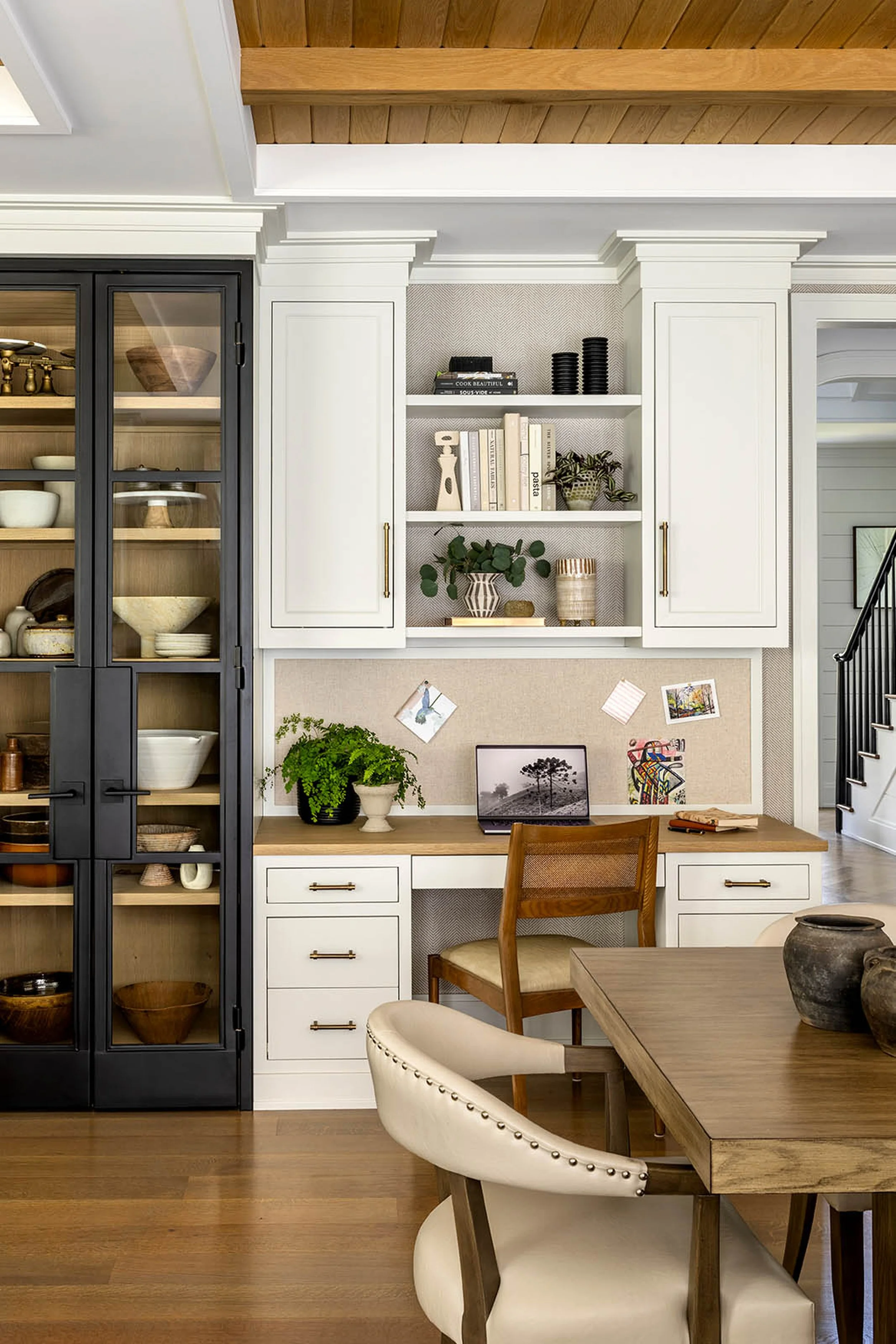 Interior view of a room showing a built-in white desk with drawers, a dark wooden chair, framed artwork, and white shelving with decorative items and books, with a wooden ceiling and hardwood floor.
