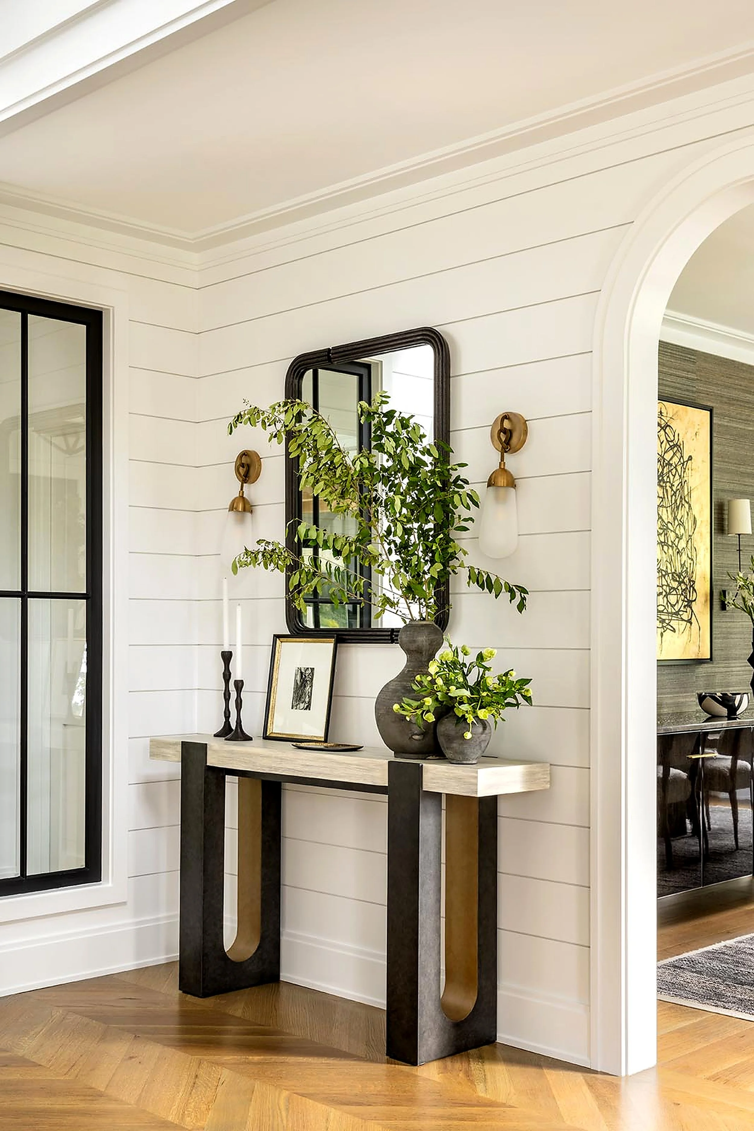 Modern console table with decorative items and vases, green plants, mirror, and framed artwork in a bright living room with white shiplap walls and arched doorway.