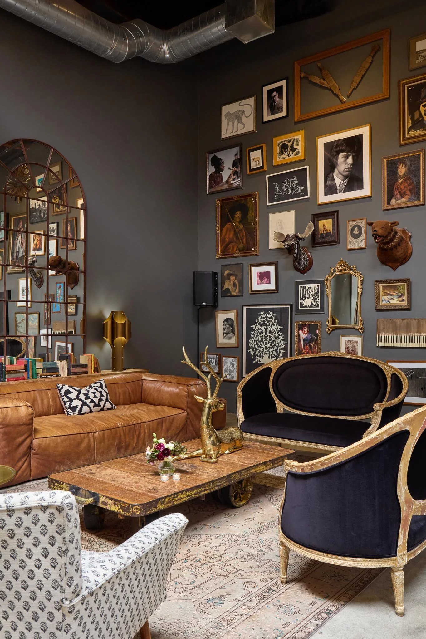 A living room with a gallery wall of framed art and photographs, including animal head mounts, including deer and moose, in a modern vintage style. There are two armchairs, a brown leather sofa, and a wooden coffee table with small candles and a flower arrangement. A gold-colored deer sculpture stands on the coffee table. The room has a gray wall and decorative area rug, with a mirror and various decorative items on the wall.
