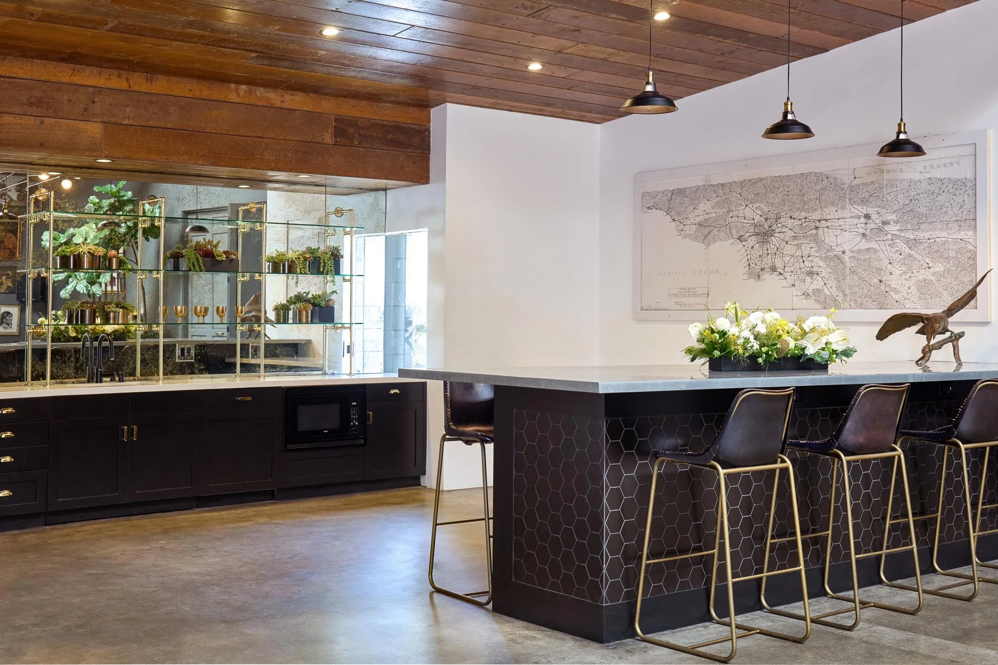 Modern kitchen with dark cabinetry, a marble island with barstools, and a wooden ceiling. There are decorative plants and a vintage bird sculpture on the counter, with a large map on the wall.