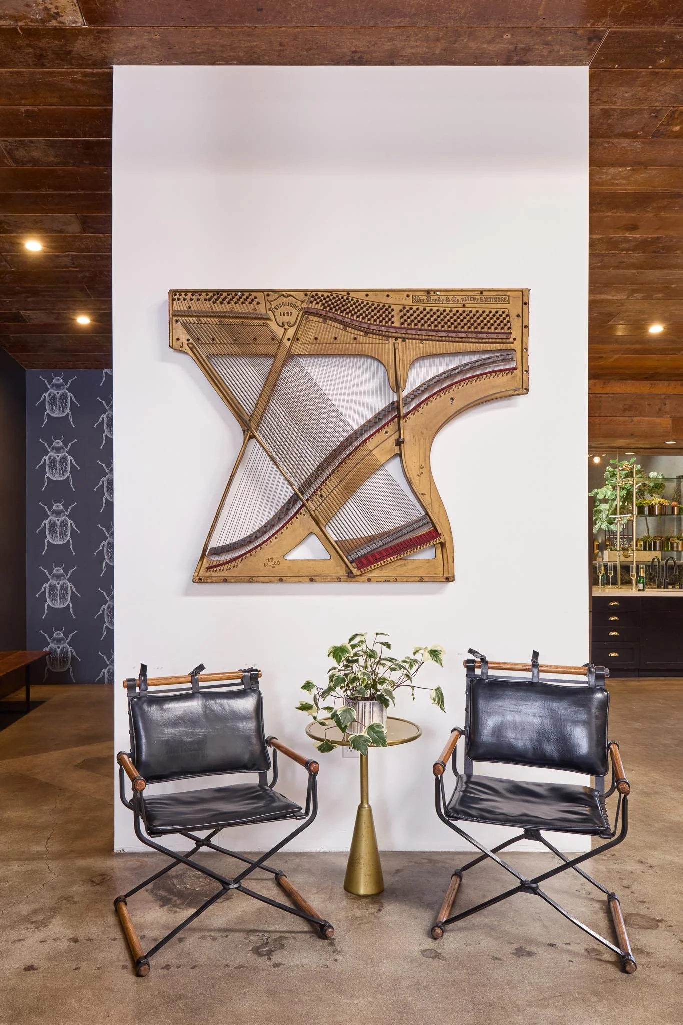 Two black leather chairs with wooden armrests facing each other, with a round table between them holding a potted green plant, against a white wall with a mounted vintage wooden piano inside a modern interior space.