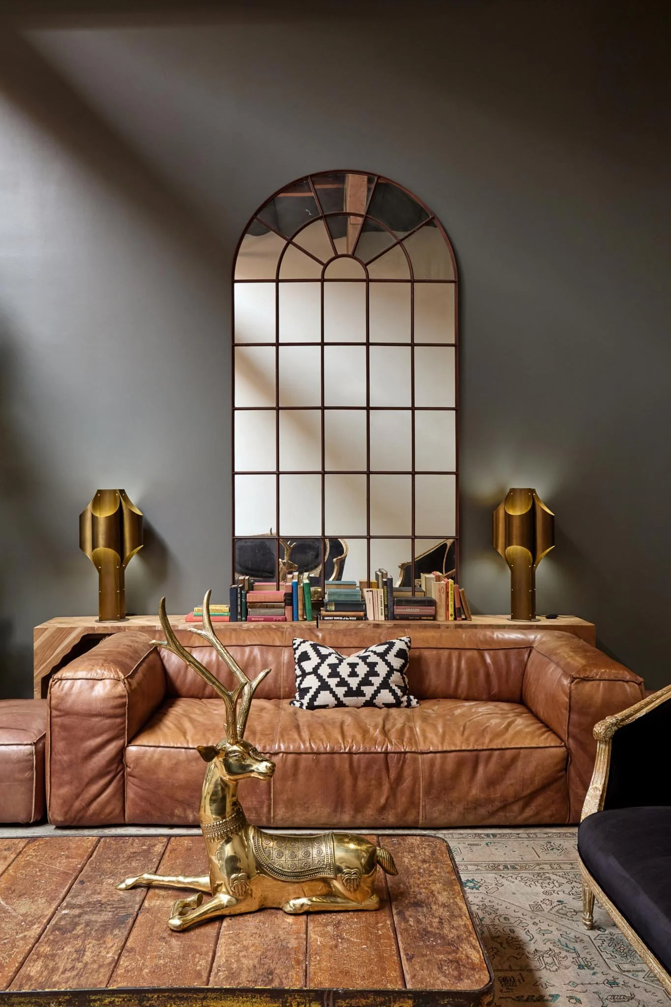 Living room with a large mirror, two gold lamps, a leather couch with a black and white patterned pillow, decorative gold reindeer sculpture on the coffee table, books behind the couch, and a patterned rug on a wooden floor.