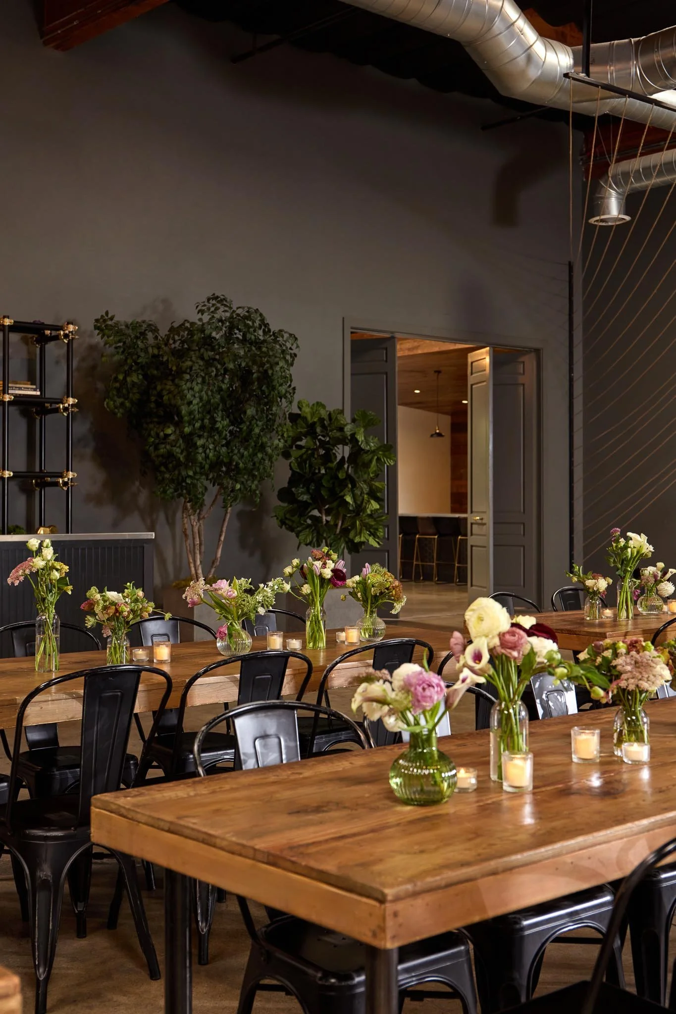 Interior of a modern event space with a long wooden table decorated with vases of pink, white, and purple flowers and small candles. In the background, there are green plants near a doorway, and the walls are gray with a decorative stripe pattern. Th