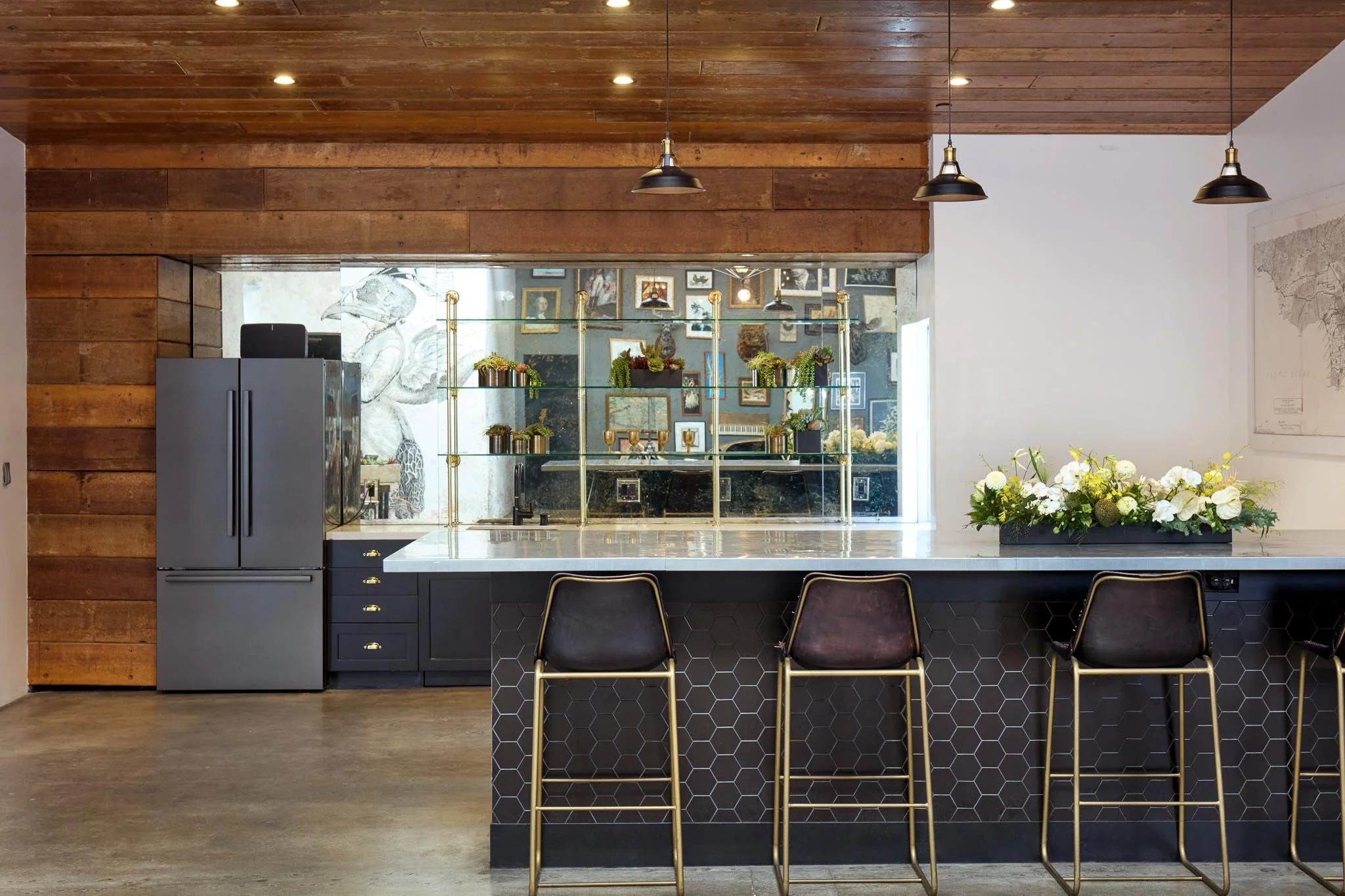 Modern kitchen with dark lower cabinets, a white marble island, and a row of black and gold barstools. Wooden ceiling and wall accents, and a floral arrangement on the island.