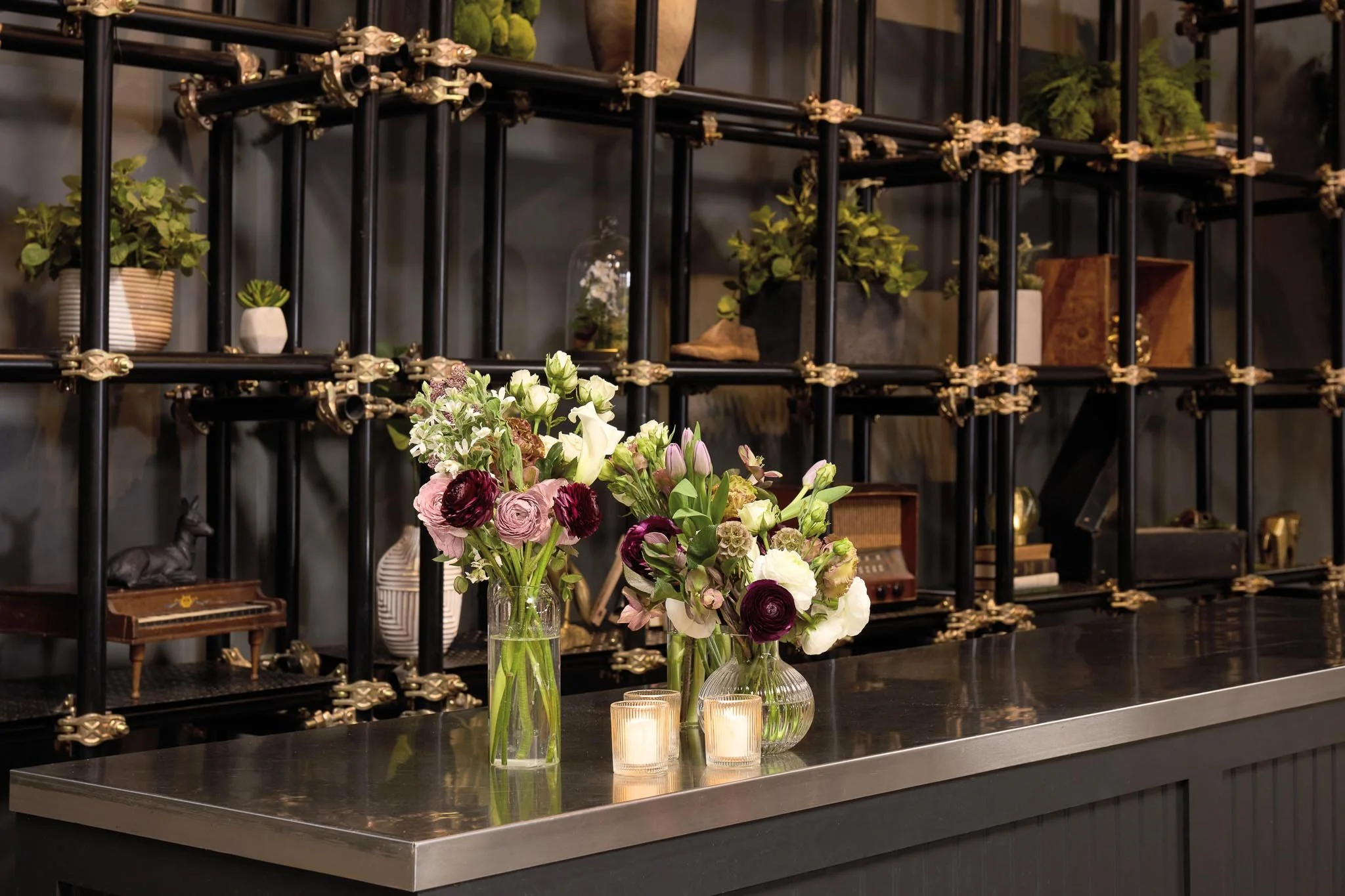Vase with mixed flowers, surrounded by candles, on a dark countertop in front of a black metal and wood shelving unit with plants and decor.