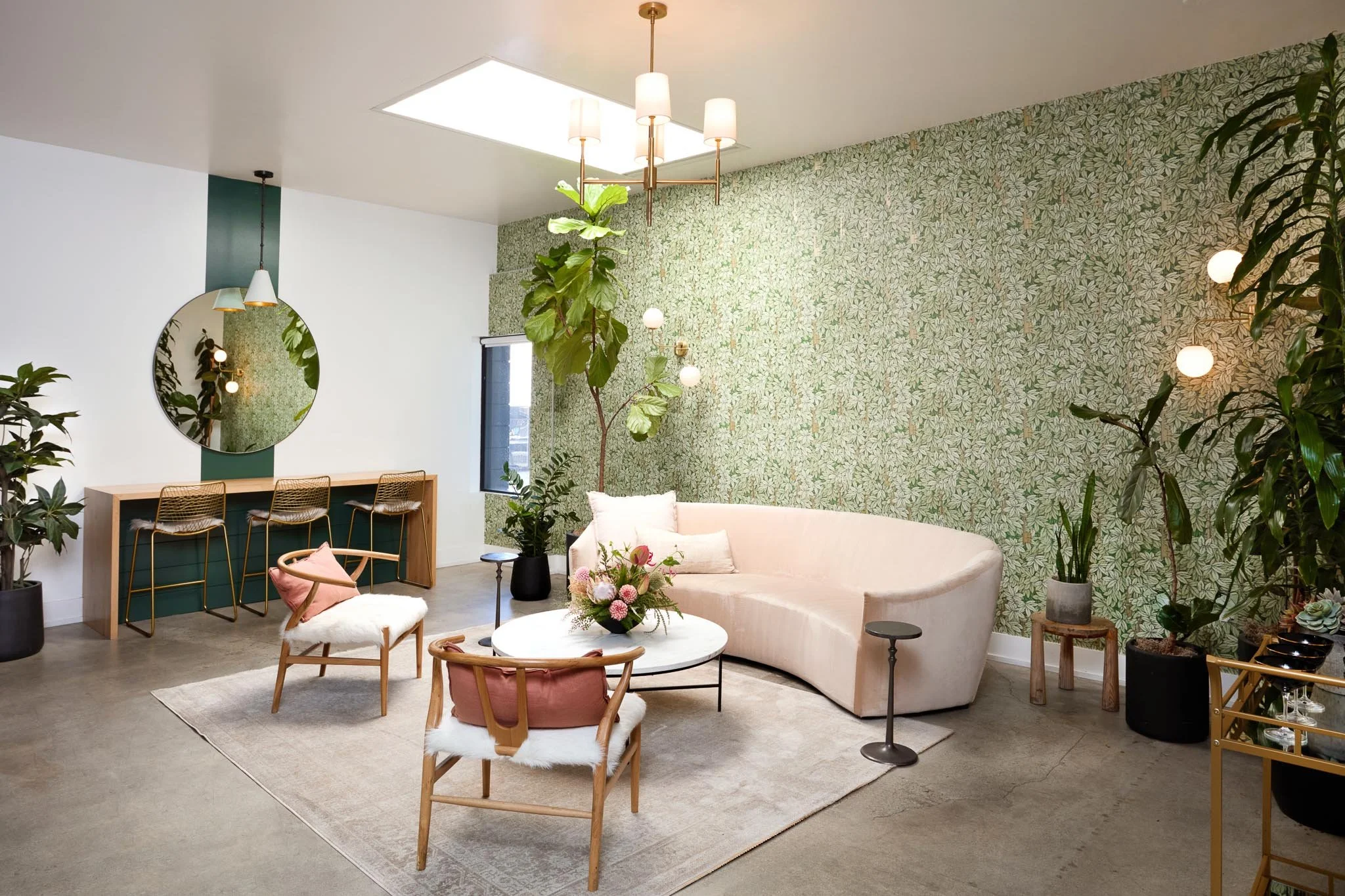 Living room with a white curved sofa, wooden chairs with sheepskin cushions, a round marble coffee table with a flower arrangement, green leafy plants, a wall with floral wallpaper, a circular mirror, a chandelier with four lamps, and a skylight in t