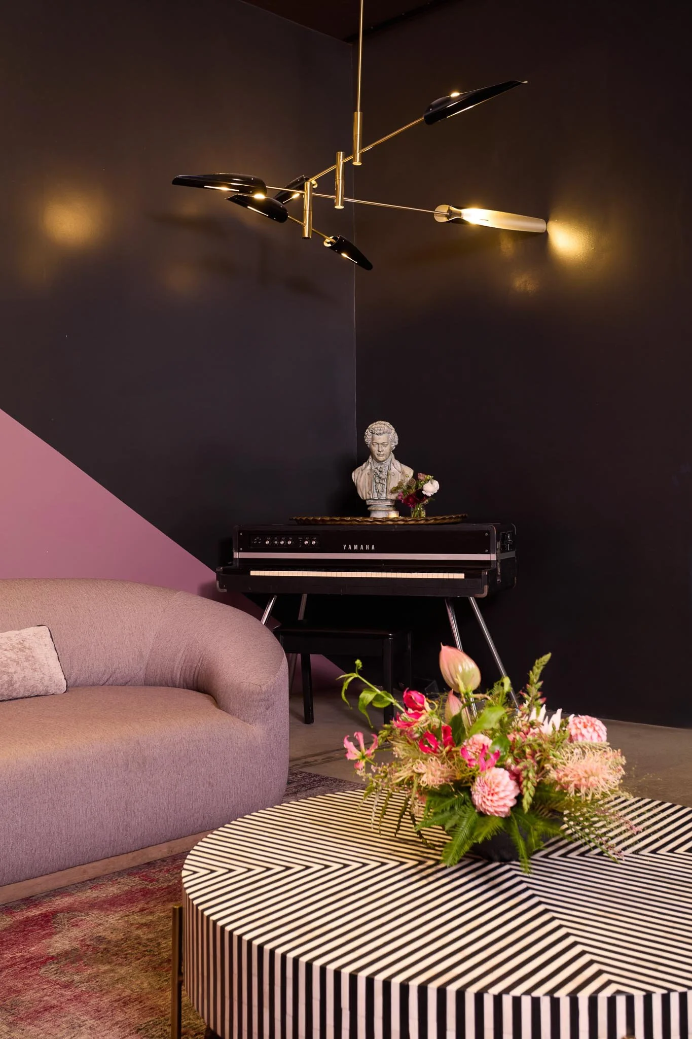 A black and white striped coffee table with a large flower arrangement, a purple sofa with a gray pillow, a black digital piano with a bust of a woman on top, and a modern ceiling light fixture against a two-tone dark wall.