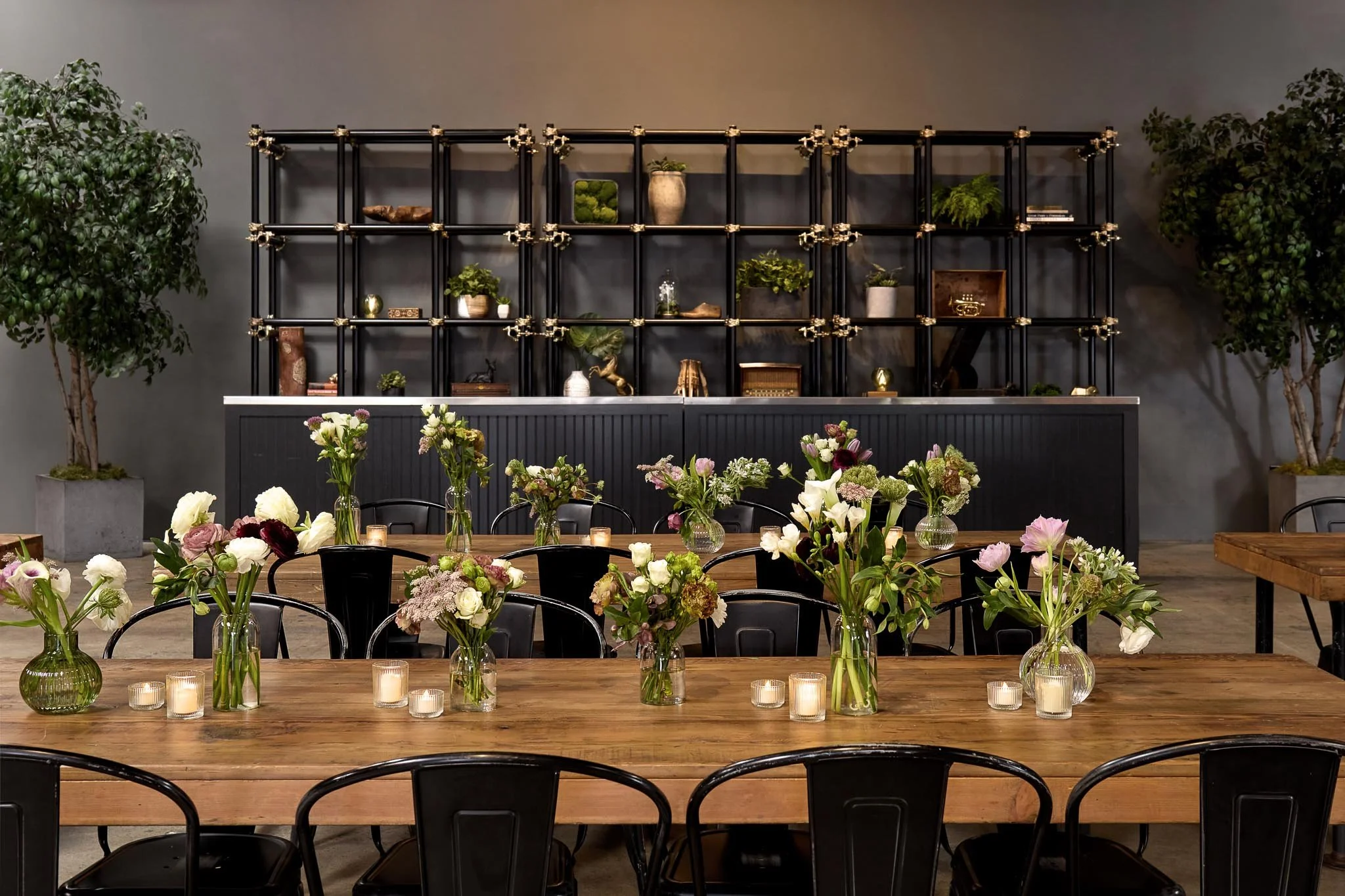 Interior view of a decorated dining space with a large wooden table, glass vases with flowers, and tealight candles. Behind the table, there is a black shelving unit decorated with plants and objects, with two large potted trees on either side. The w