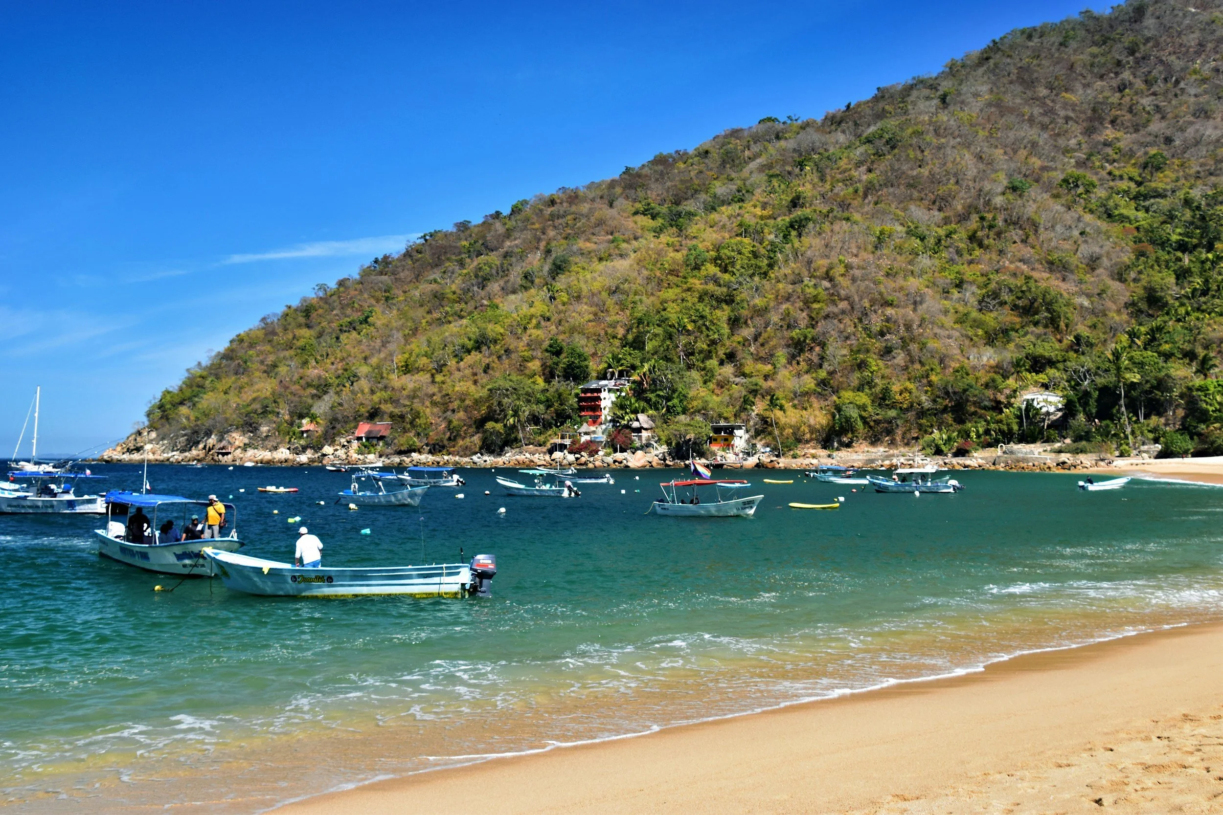 A scenic beach with light brown sand and clear turquoise water, with boats anchored near the shore. In the background, a hillside covered with green and brown vegetation rises above the water, with small buildings nestled at its base. The sky is bright blue.