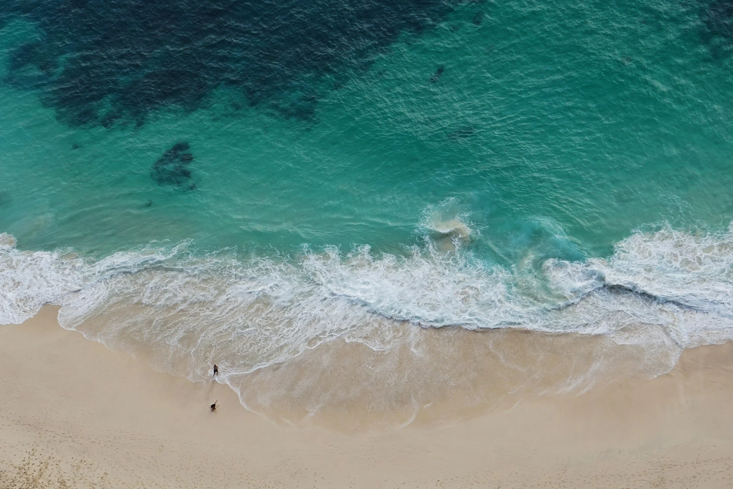An aerial view of a sandy beach with waves washing ashore and a person standing in the water.