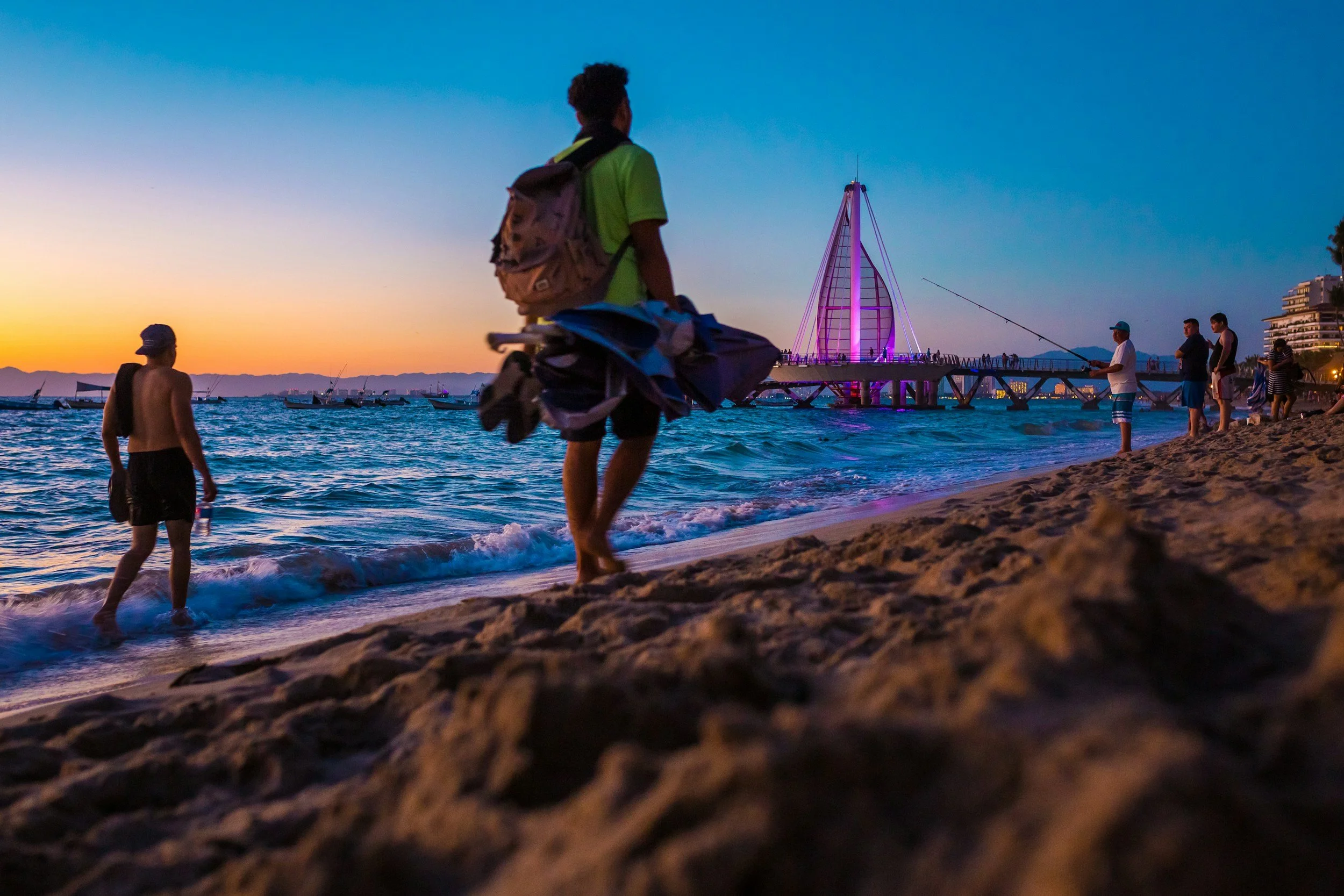 People fishing on a beach during sunset, with a lit-up pier and boats in the water.