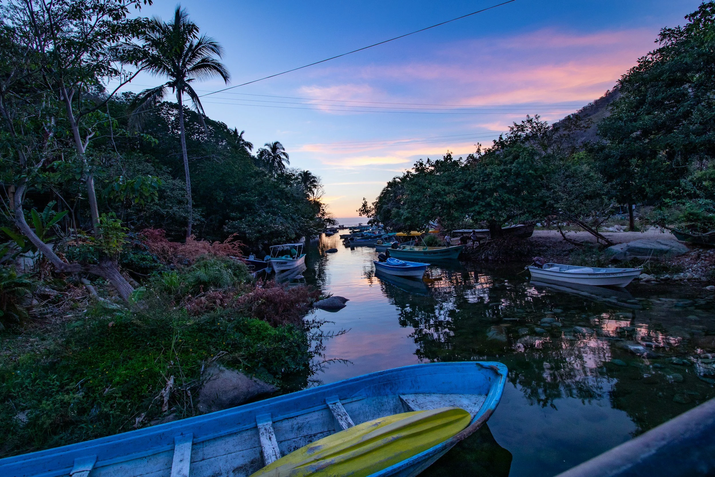 A serene view of a small boat on a calm waterway at sunset, surrounded by lush green trees and hills, with a purple and pink sky.