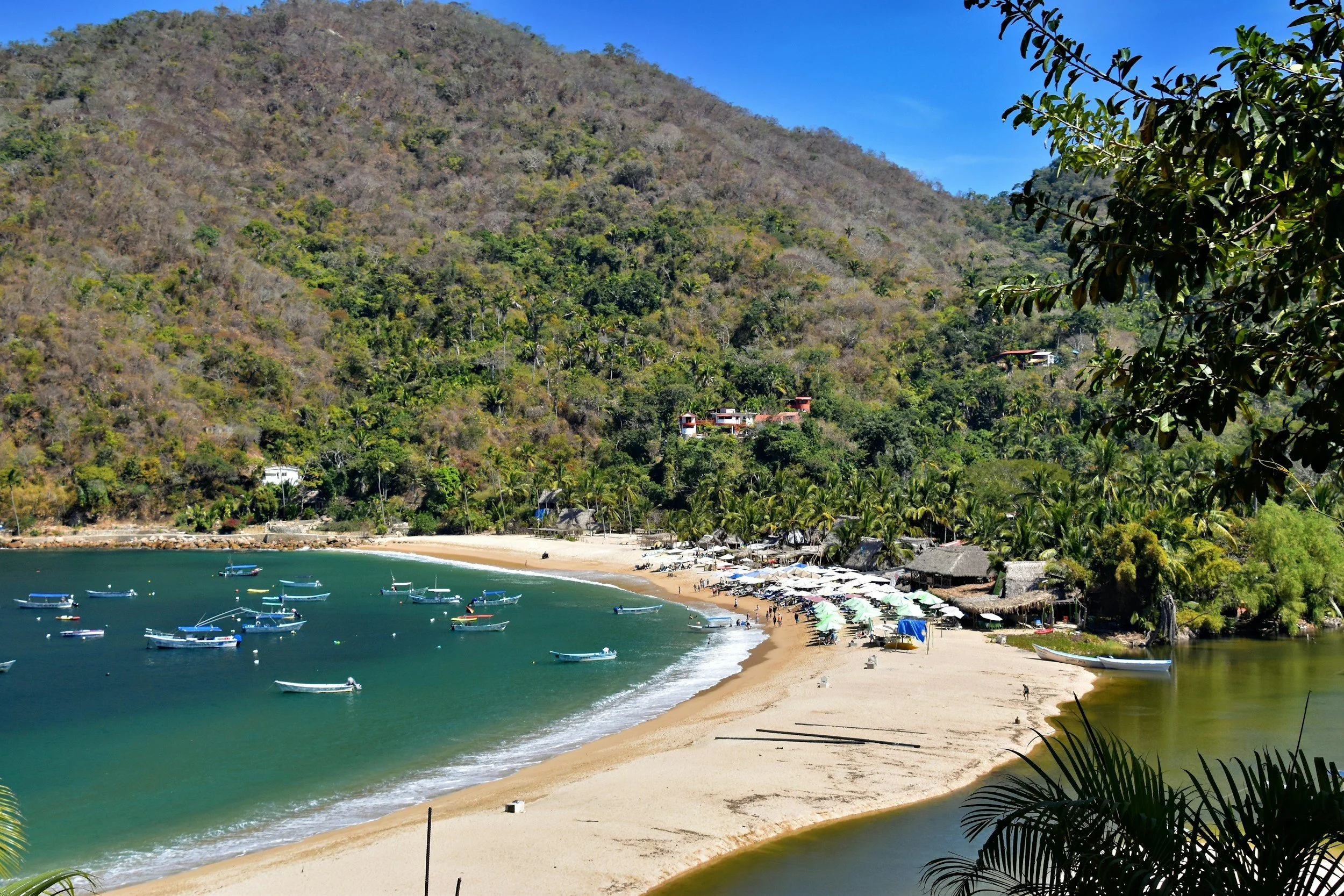 A tropical beach with white sand, surrounded by lush green hills and clear blue water. There are many boats anchored near the shore and beach umbrellas and lounge chairs on the sand. The sky is clear and blue.
