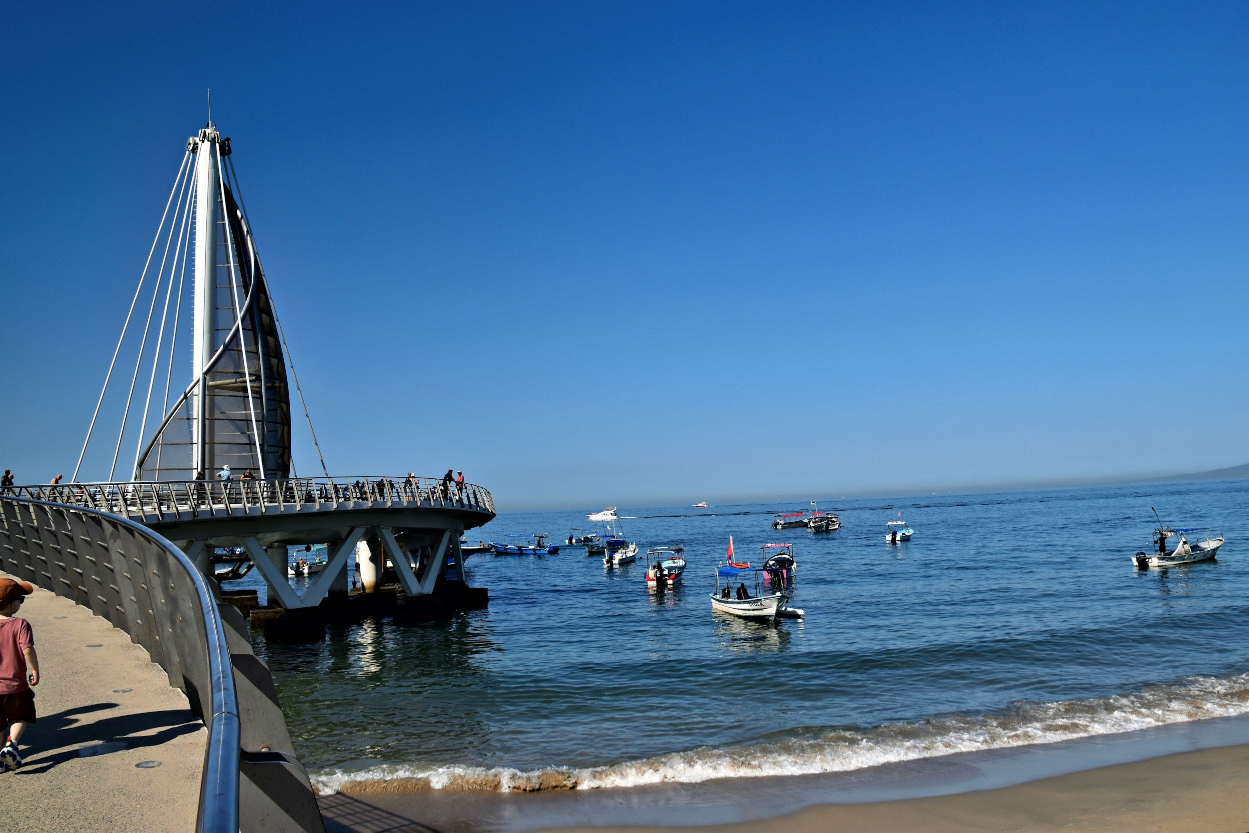 A pier with a modern sail structure extending into a marina filled with boats, with a clear blue sky and calm sea in the background.