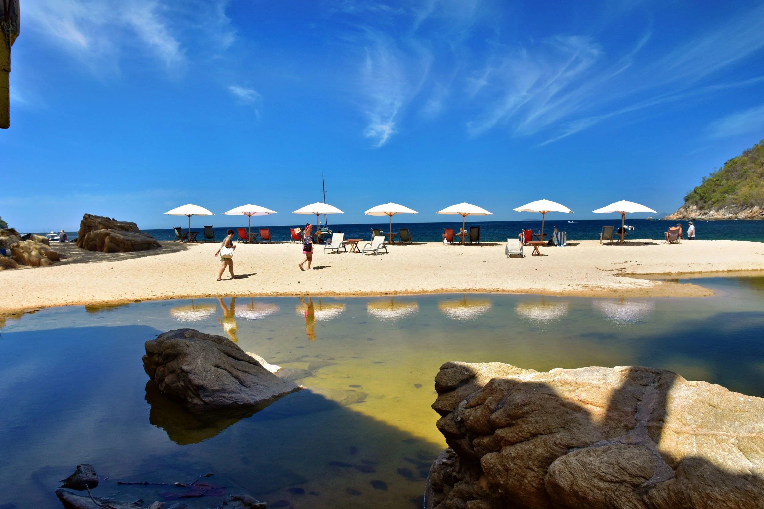 A beach scene with white umbrellas and lounge chairs, people walking, rocky foreground, and a hillside on the right under a blue sky with wispy clouds.
