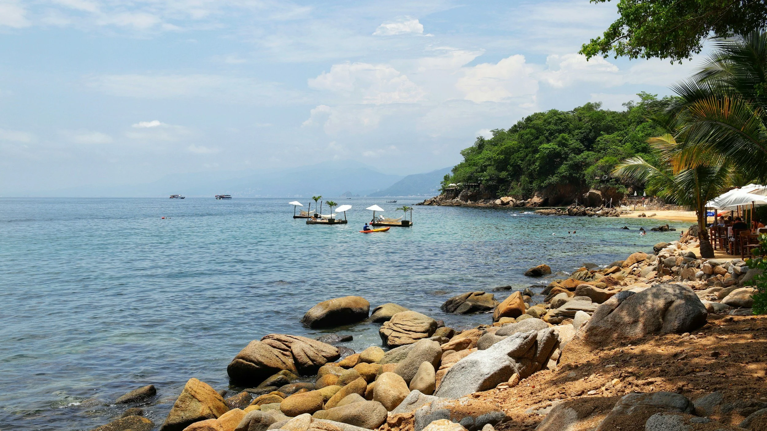 A scenic view of a tropical beach with rocky shoreline, palm trees, and a clear blue ocean. There are boats on the water and a lush, green hill in the background under a partly cloudy sky.