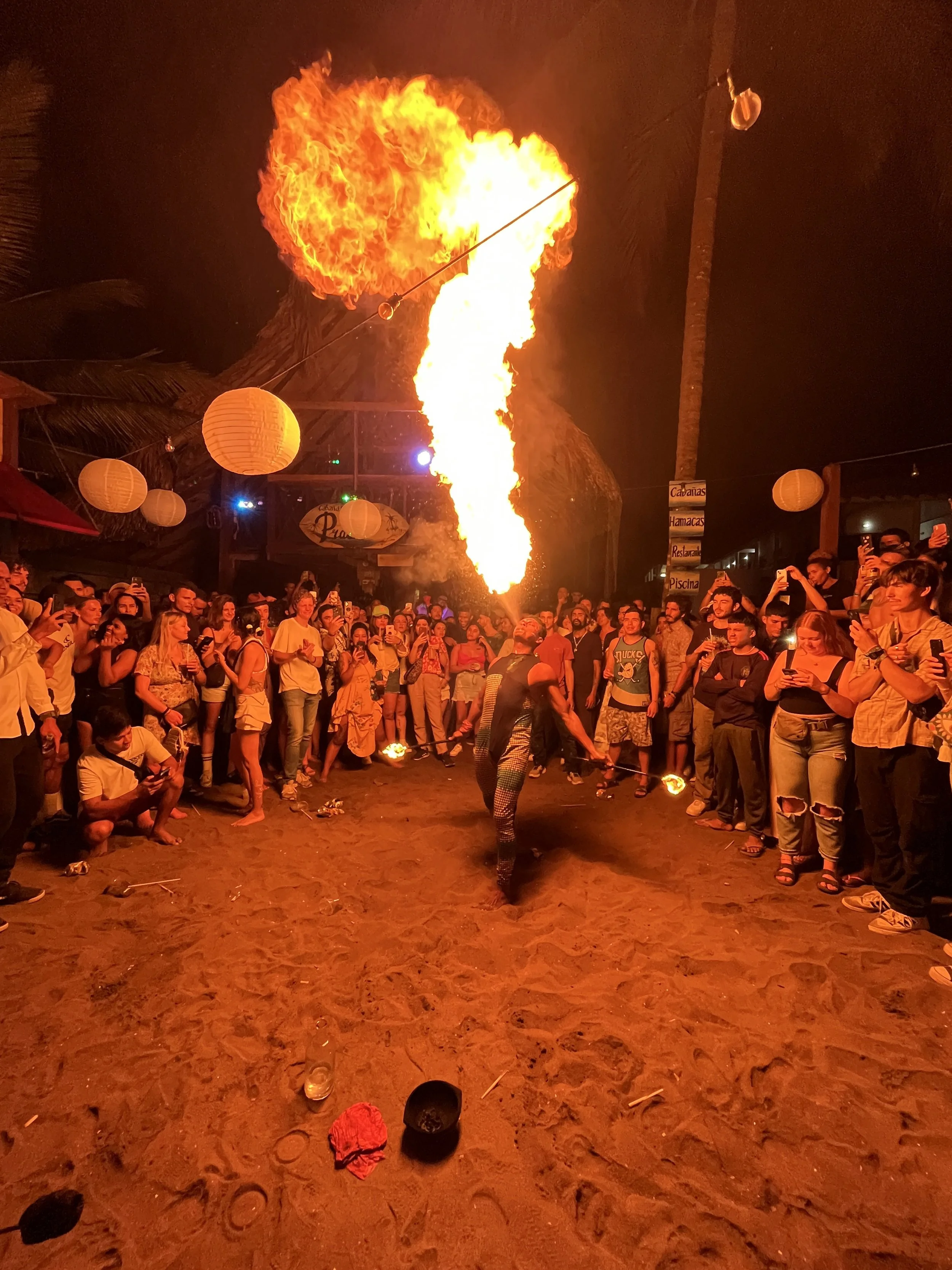 A fire performer shooting a large flame into the air during a fire show at night, with a crowd of spectators watching and taking photos on a sandy outdoor venue decorated with paper lanterns.