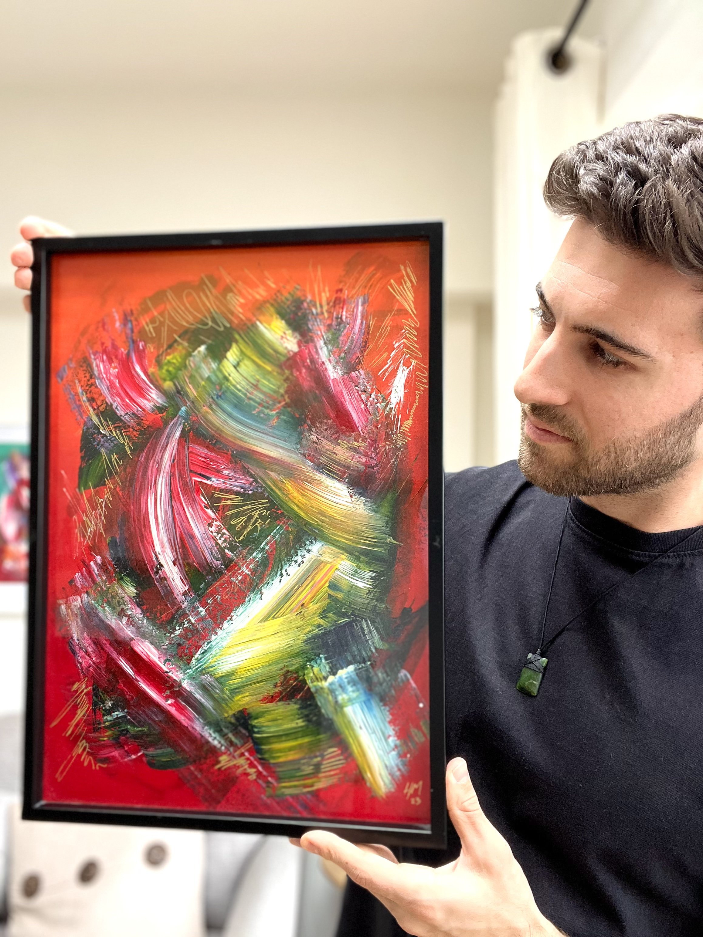A man with dark hair and short beard holding an abstract painting in a black frame, with bright red background and colorful brushstrokes of yellow, white, green, black, and pink.