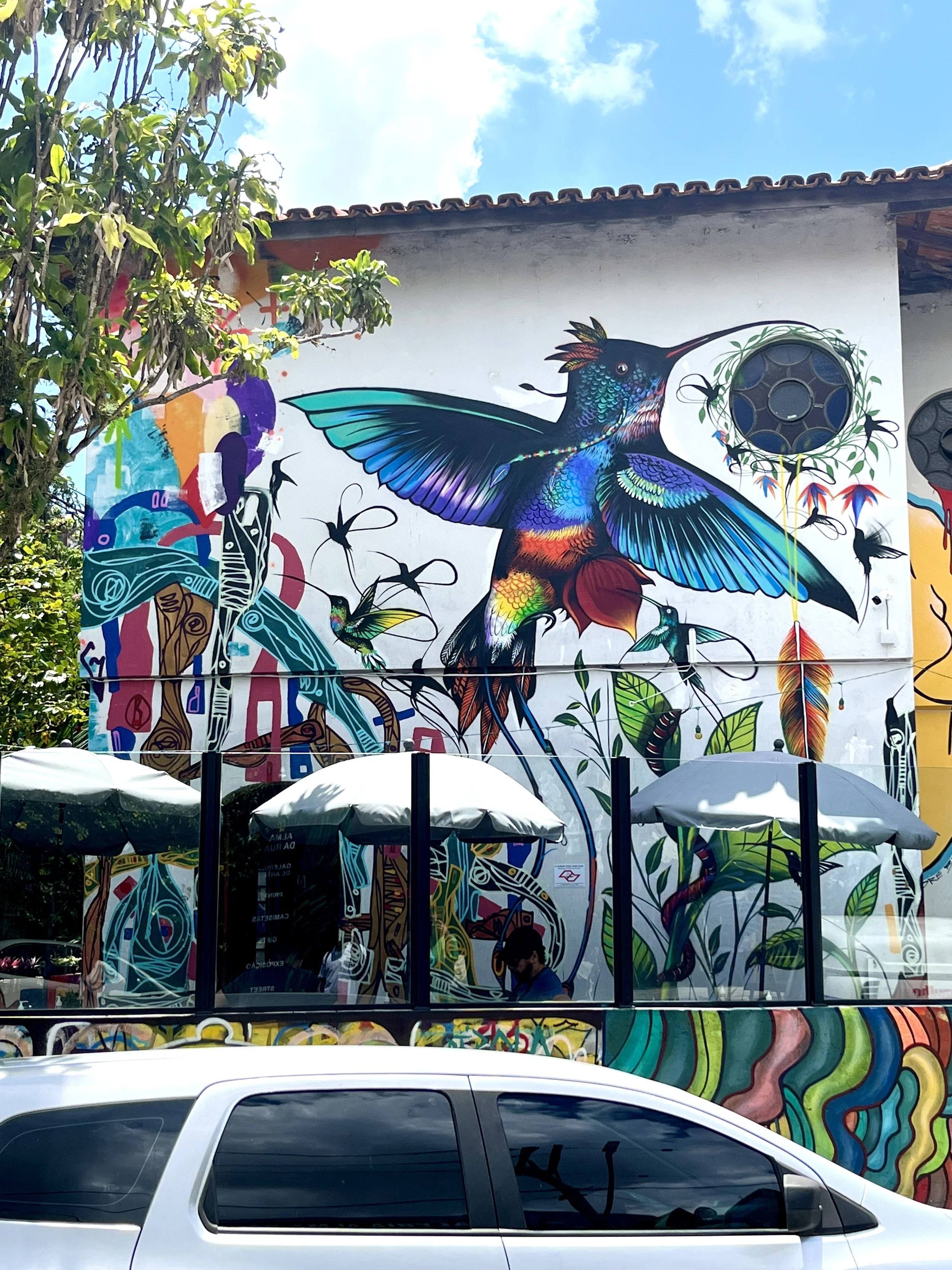 Colorful mural of a hummingbird with vibrant feathers on a white wall, surrounded by abstract plants and shapes. There are umbrellas and a person in the foreground.