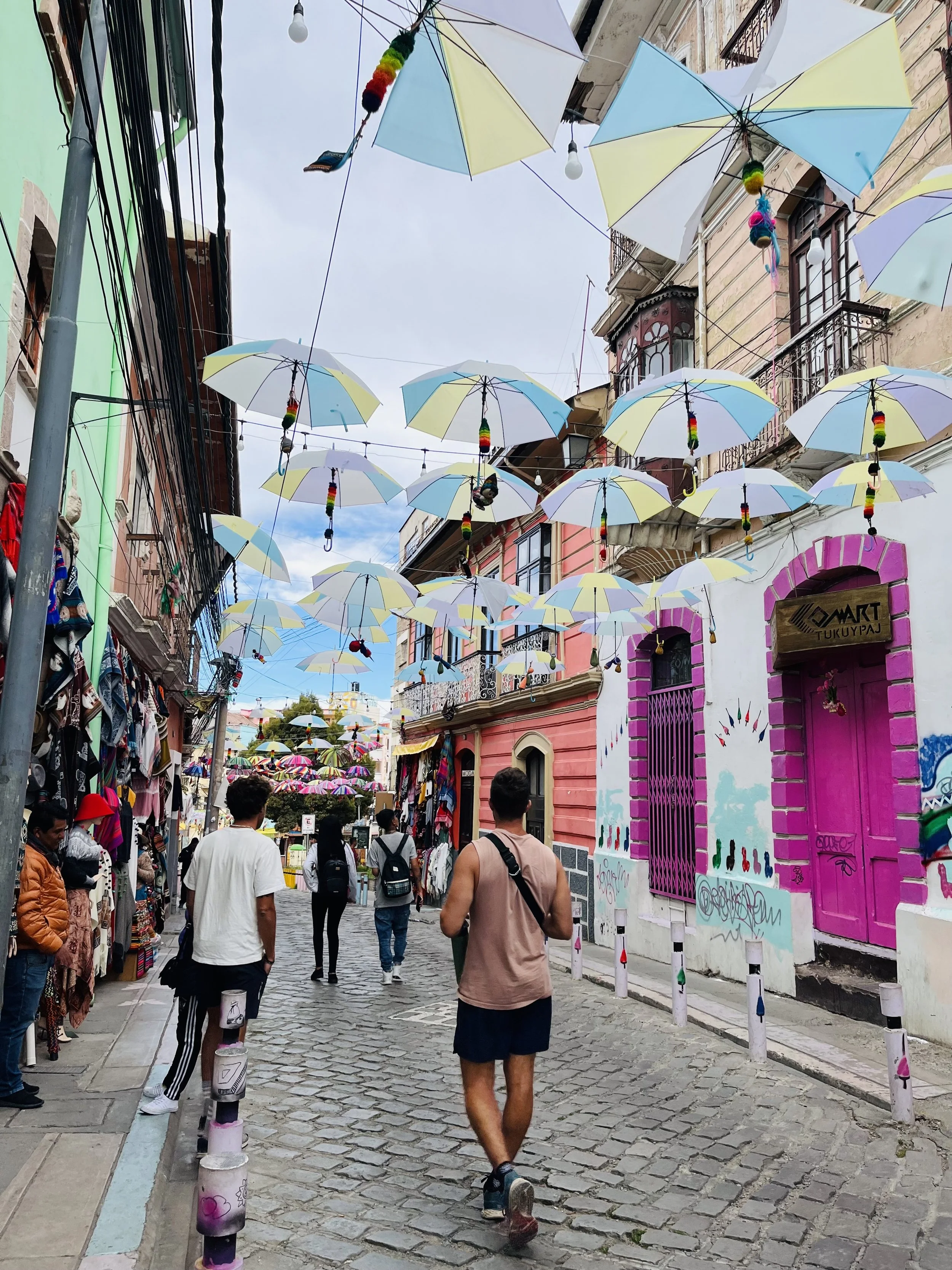 Colorful umbrellas hanging above a cobblestone street with people walking, shops with vibrant storefronts, and buildings with European-style architecture.