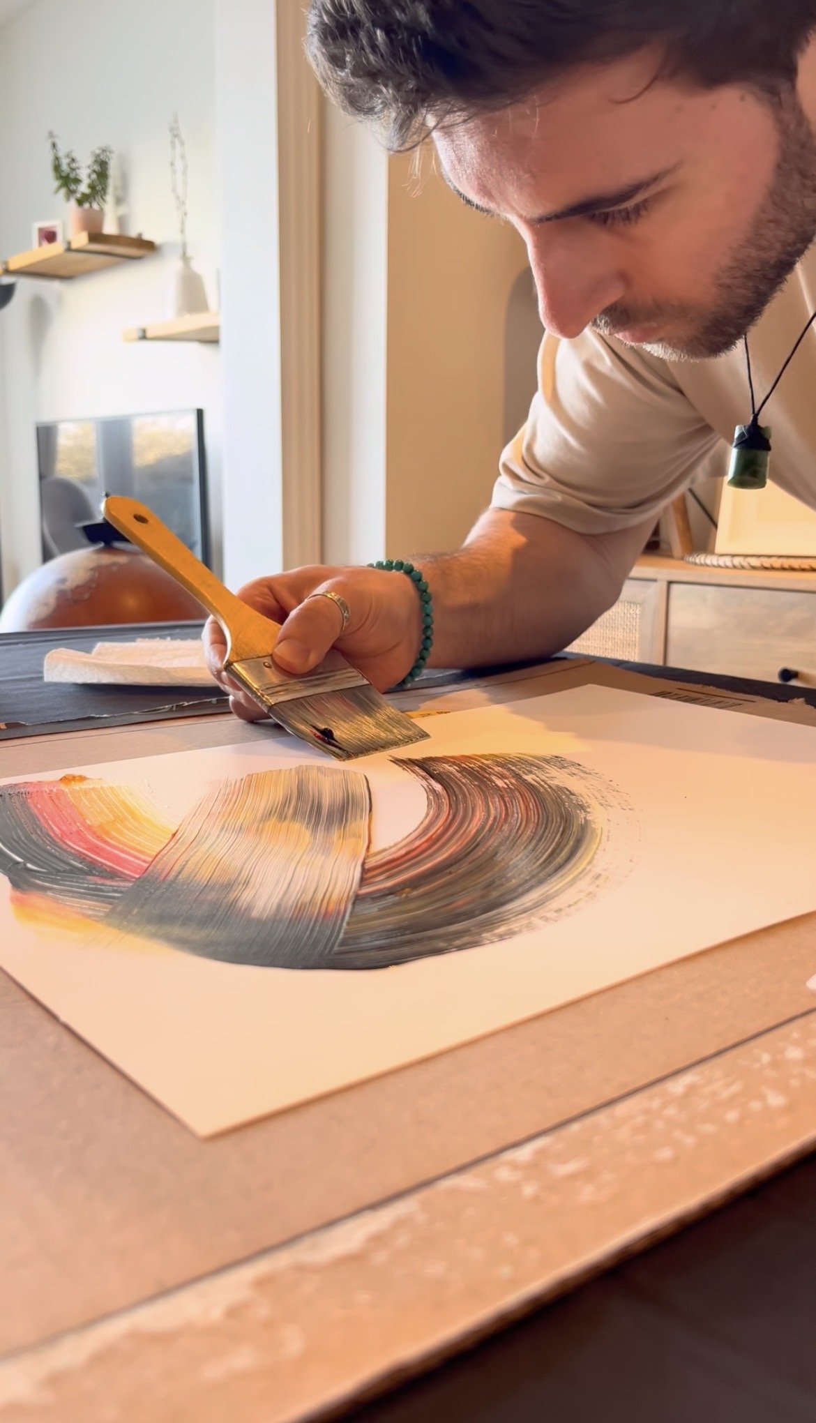 A man painting with a broad brush on a canvas, creating an abstract swirl of colors including yellow, red, gray, and black, inside a well-lit room.