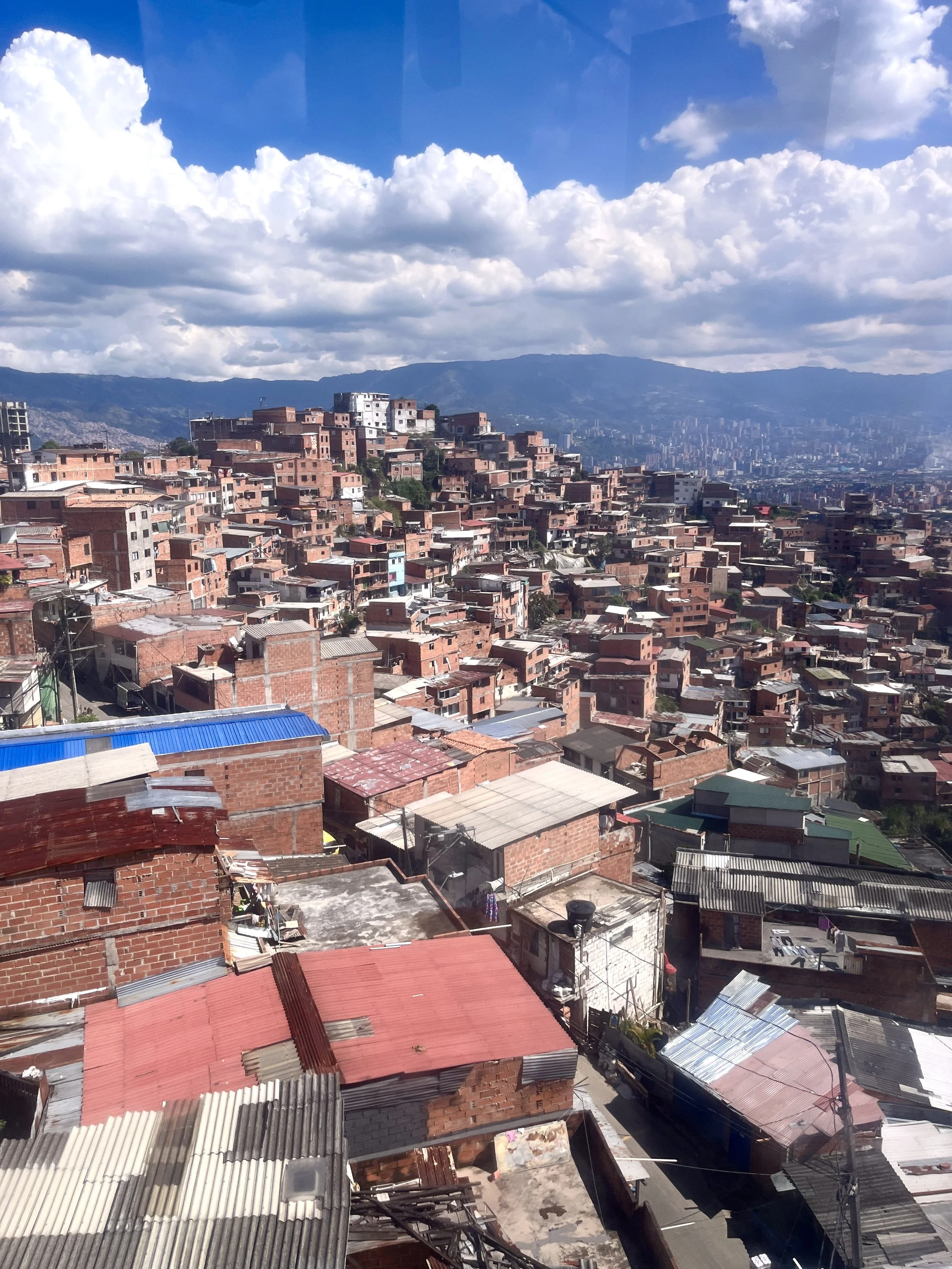 View of a hilly urban neighborhood with densely packed brick buildings and colorful rooftops, under a partly cloudy sky with mountains in the background.