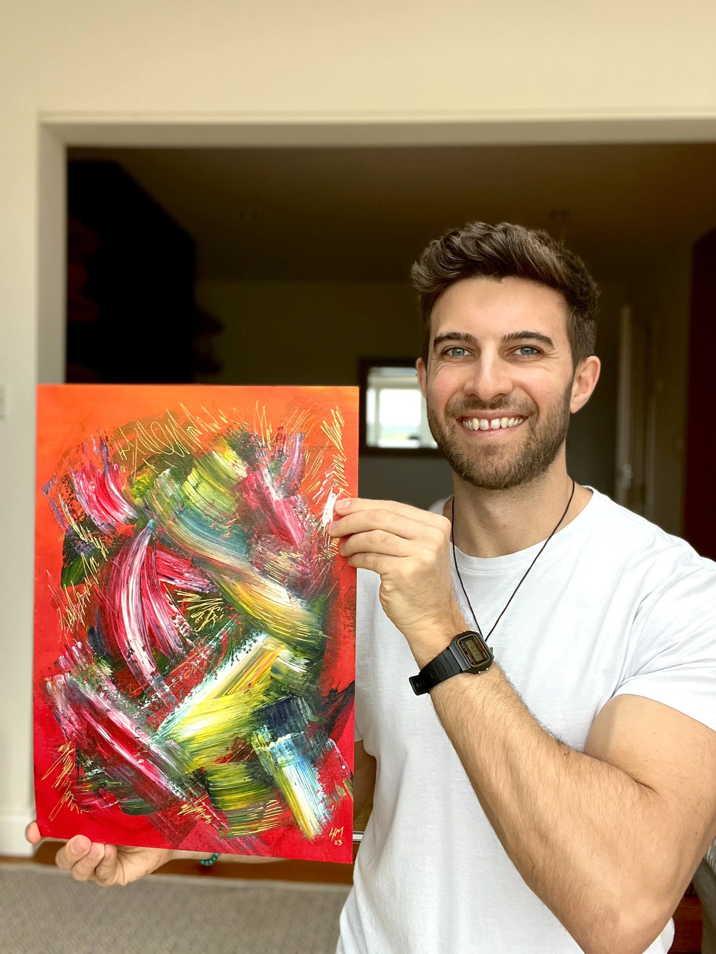 A smiling man with a short beard and dark hair holding an abstract colorful painting on a red canvas.