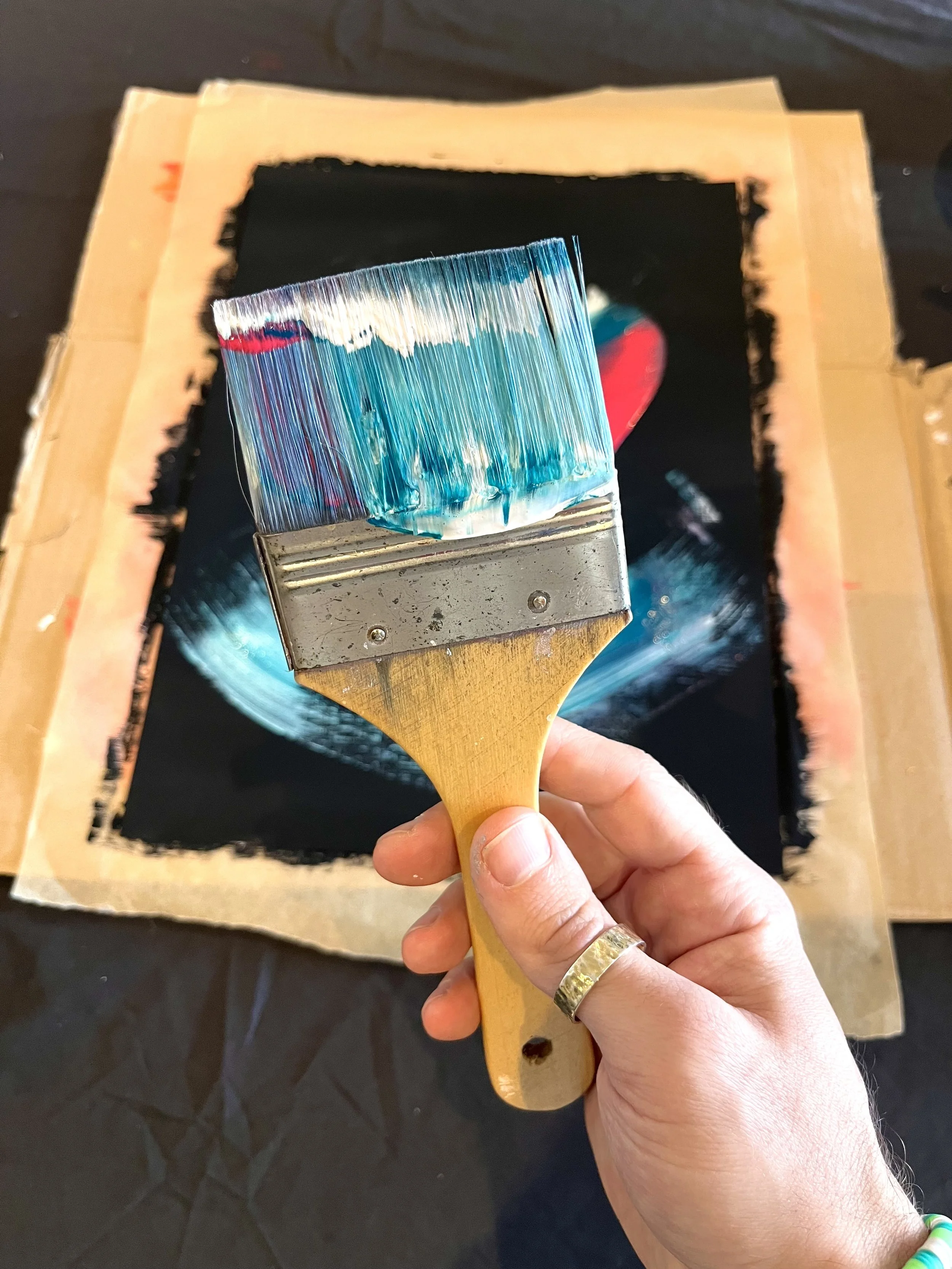 Person holding a paintbrush dipped in blue and white acrylic paint, preparing to paint on a black canvas, with masking tape around the edges.