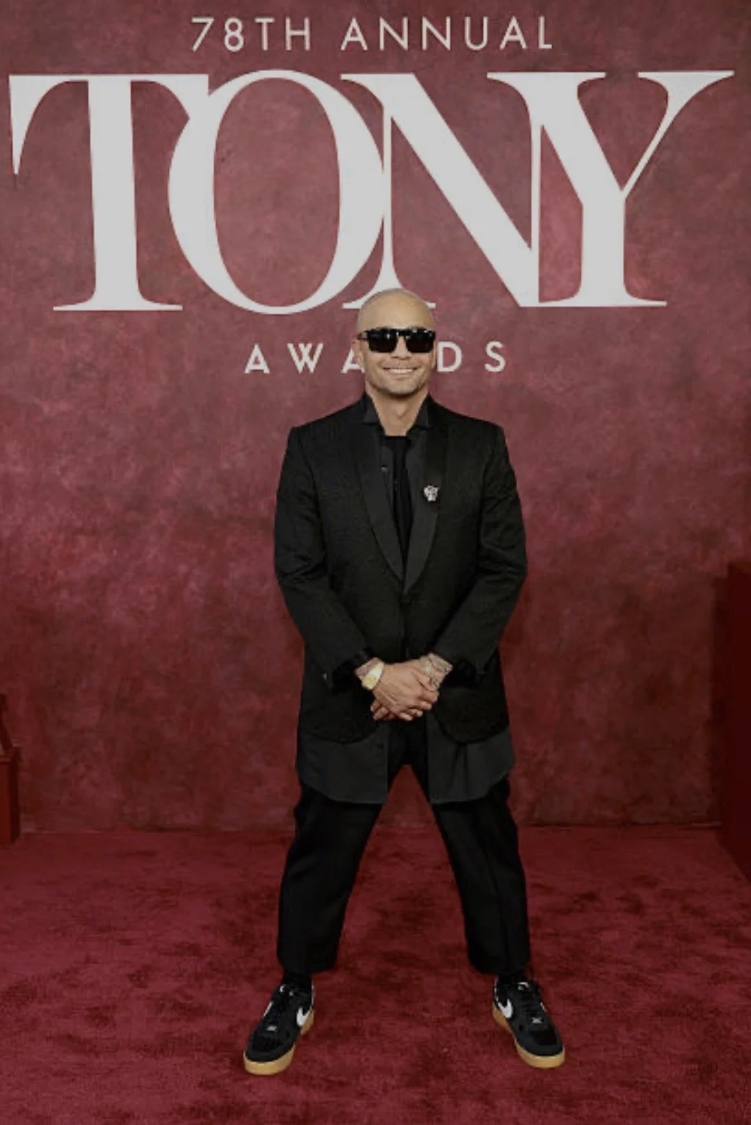 Seth Stewart standing on a red carpet at the 78th Annual Tony Awards, wearing sunglasses, a black suit, and sneakers, in front of a backdrop with the Tony Awards logo.