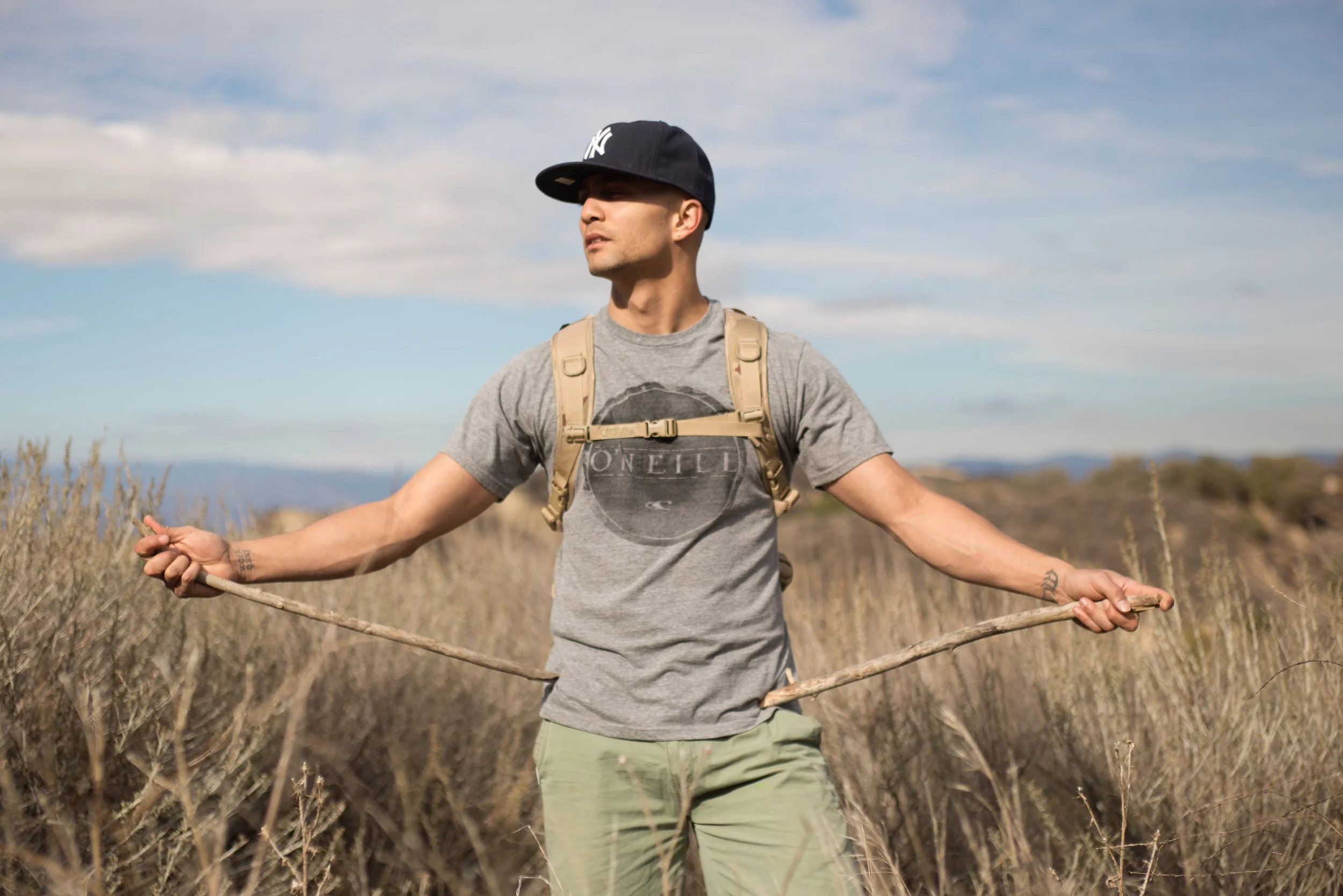 Seth Stewart, a young man standing outdoors in a field with tall grasses, wearing a black cap, gray t-shirt, and carrying a tan backpack. He holds a walking stick in each hand, with arms extended and looking to the side, under a partly cloudy sky.