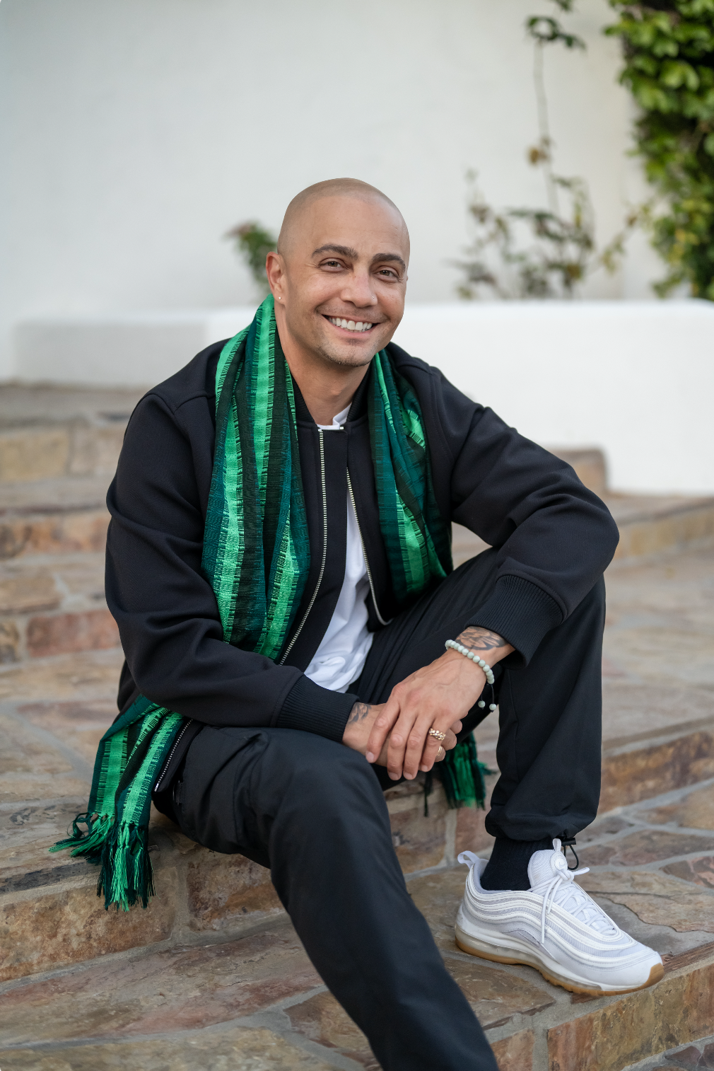 Seth Stewart, wearing a black jacket, white shirt, black pants, white sneakers, and a green patterned scarf, sitting on stone steps outdoors with plants in the background.