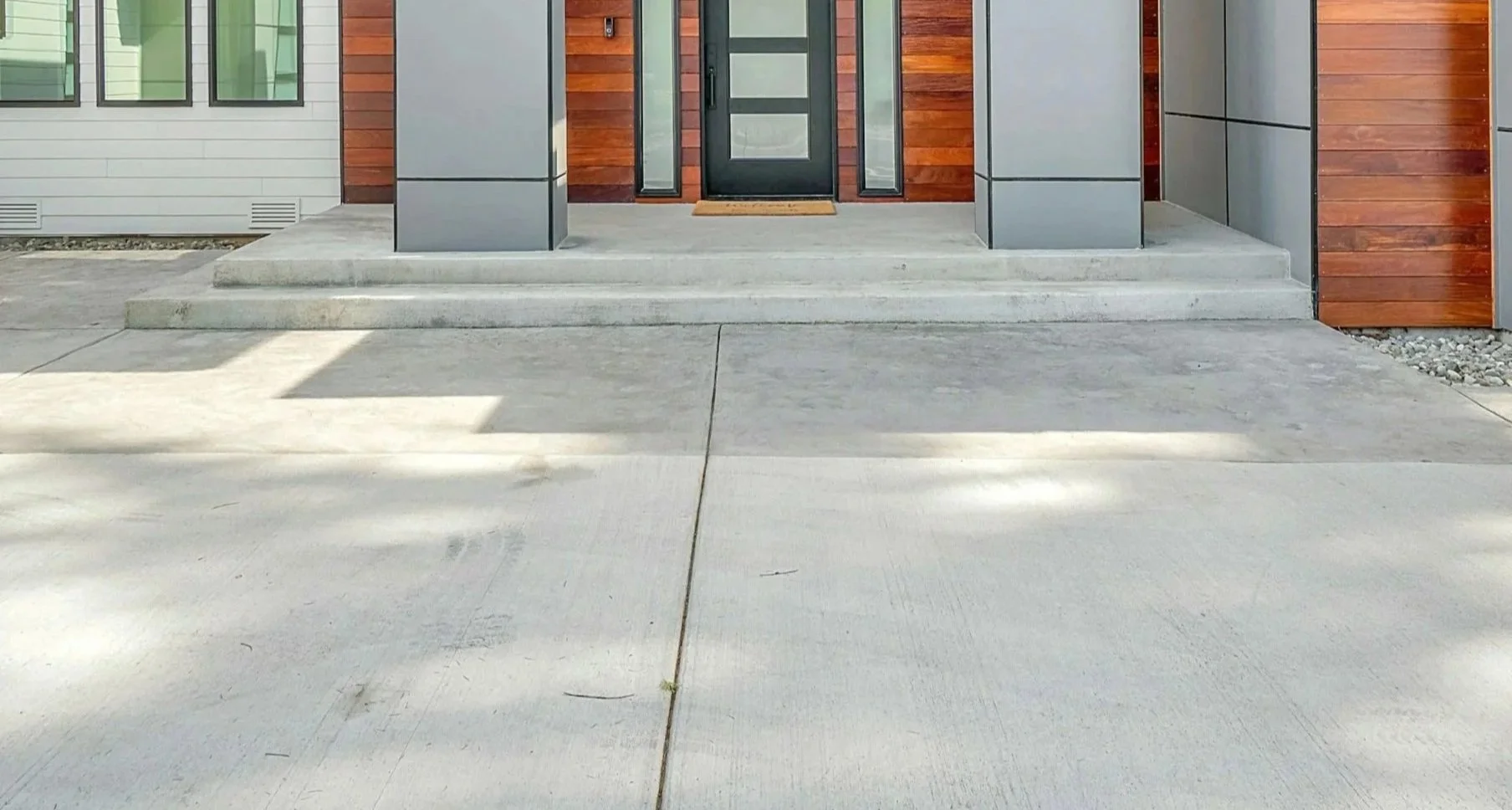 Concrete steps leading up to a modern front door with black frame and glass panel, flanked by large grey and wooden siding walls.