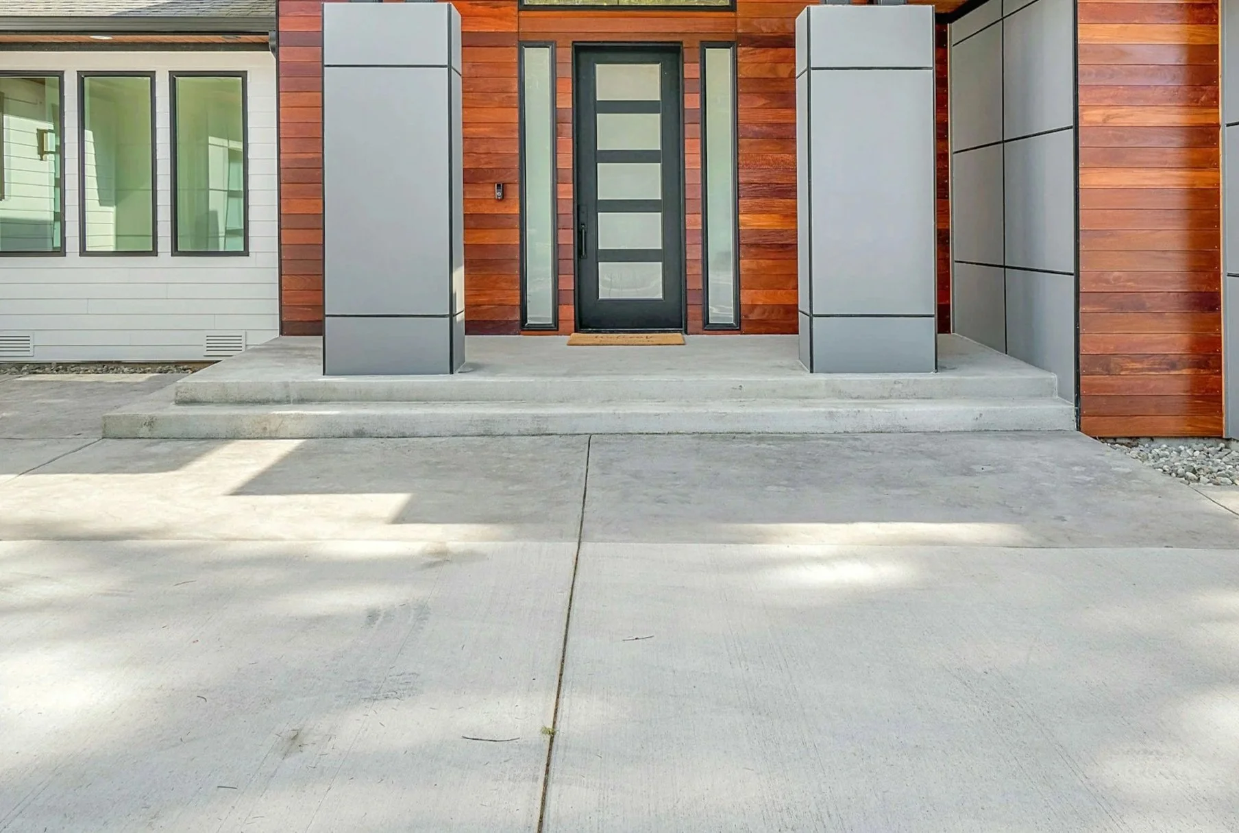 Modern house entrance with a black door, wooden siding, and large grey metal panels, flanked by narrow sidelight windows, concrete steps, and a concrete porch.