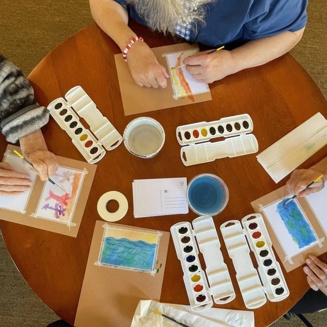 Several people sitting around a round wooden table painting watercolor pictures. There are watercolor palettes, cups of water, paper, and paintbrushes on the table.