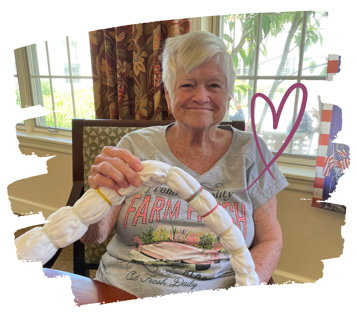An elderly woman with short white hair smiling and sitting at a table with large windows behind her. She is holding a white fabric-covered object, possibly a handcrafted item, in her hands. There is a purple heart drawing on the image.