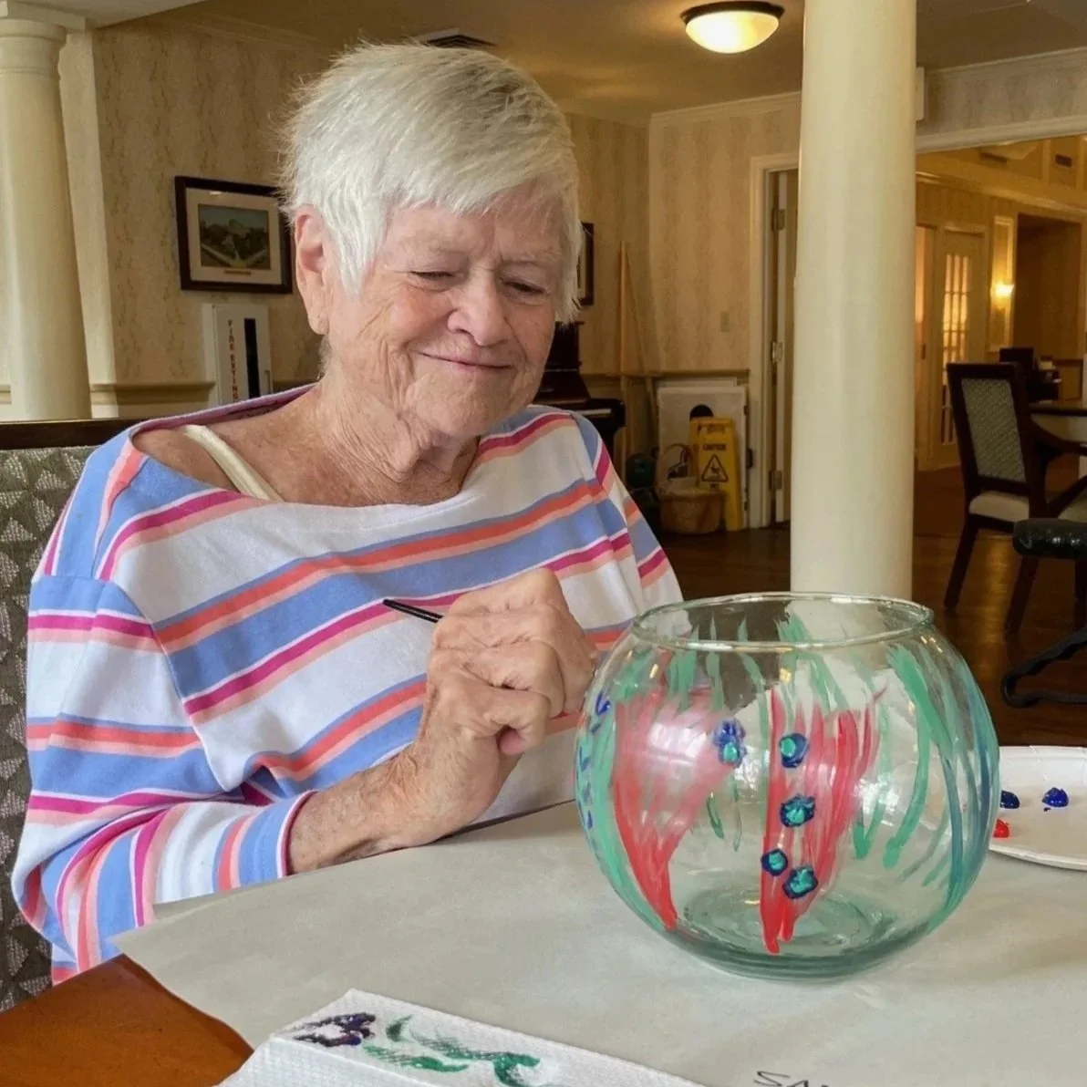 An elderly woman in a striped shirt is painting a glass bowl with red, green, and blue colors.