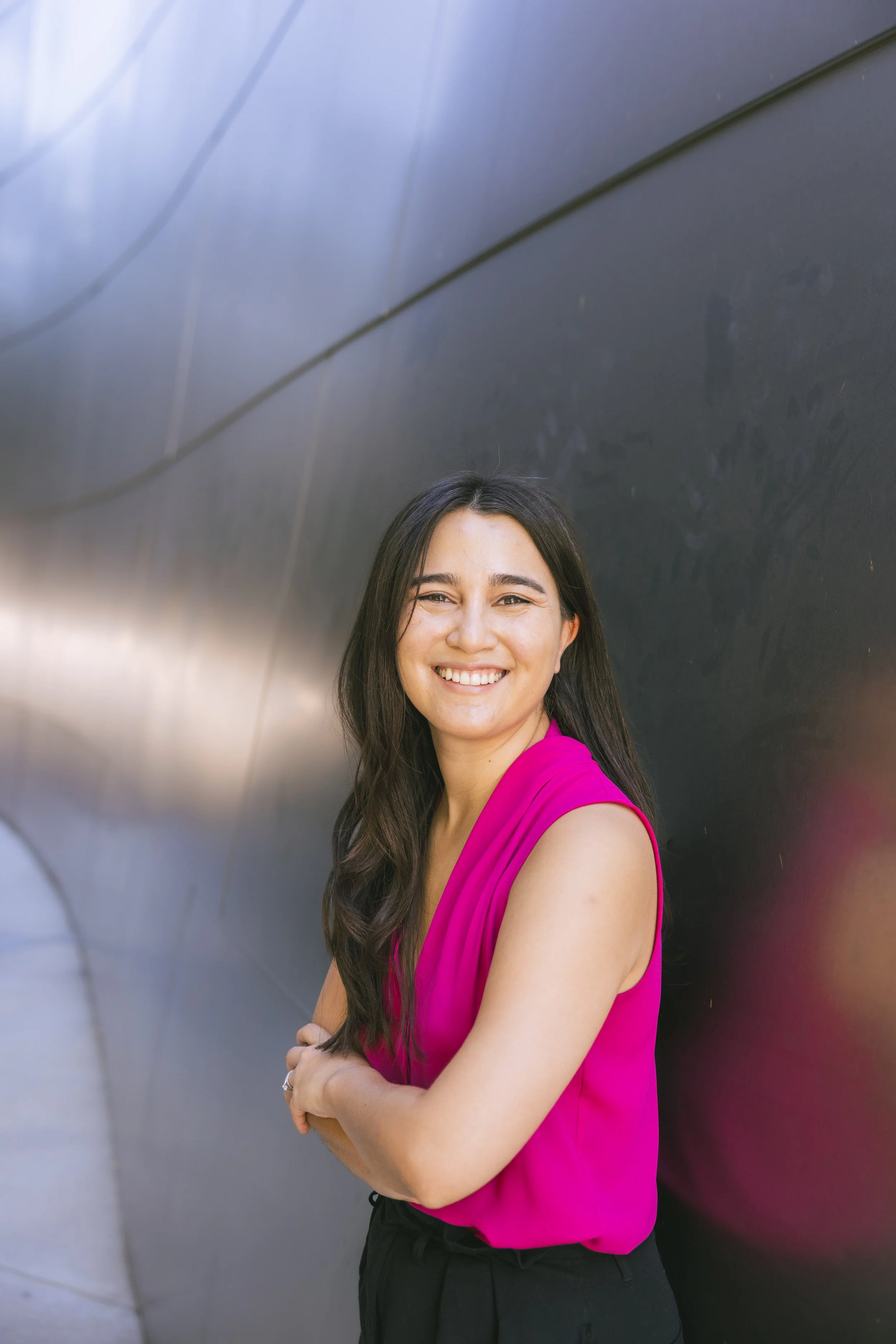 A smiling woman with long dark hair wearing a bright pink sleeveless top standing against a metallic surface background.