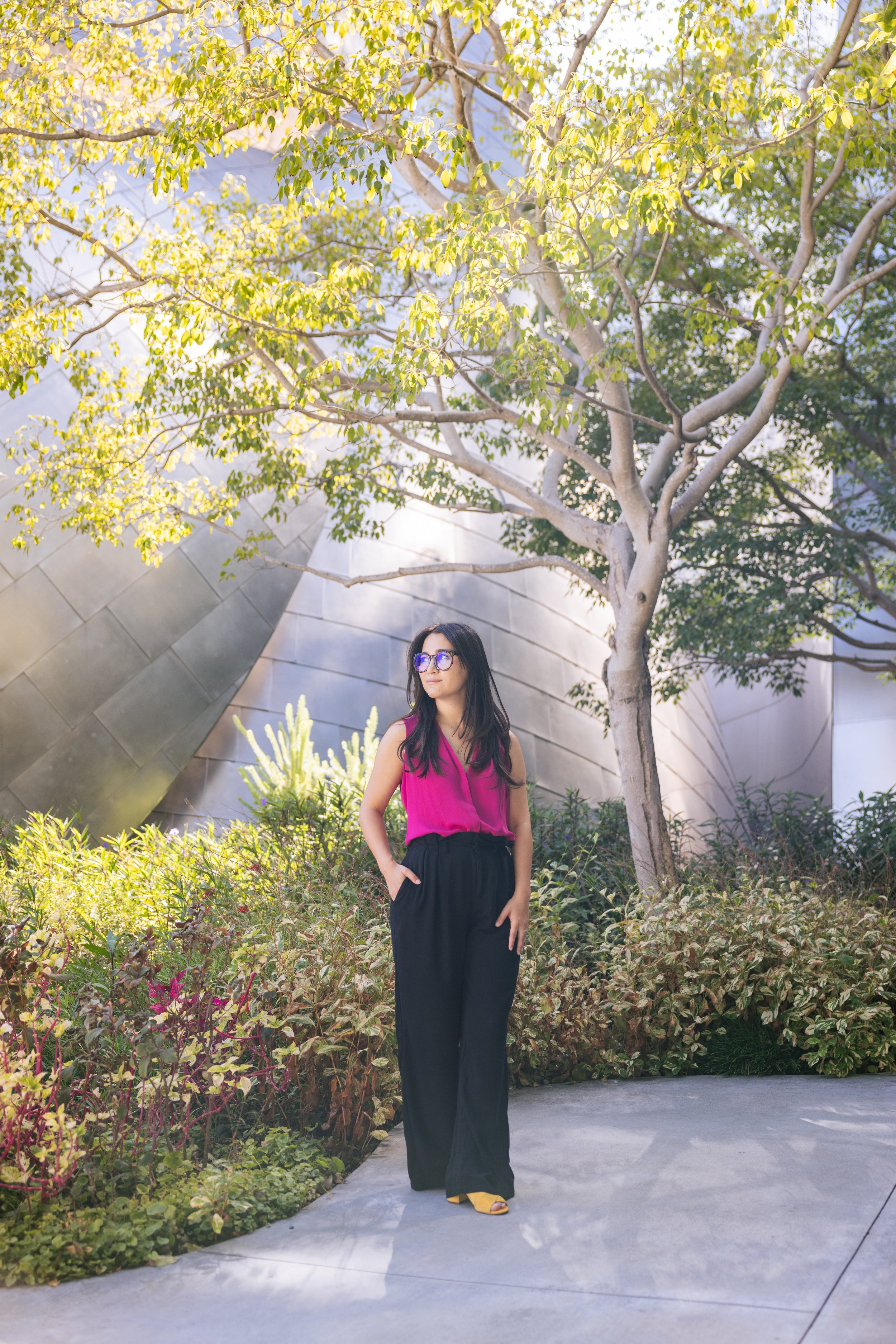 A woman with long dark hair and glasses wearing a sleeveless magenta top and black pants, standing outdoors on a paved path surrounded by greenery and yellow flowers, with a modern metallic building and a large leafy tree in the background.