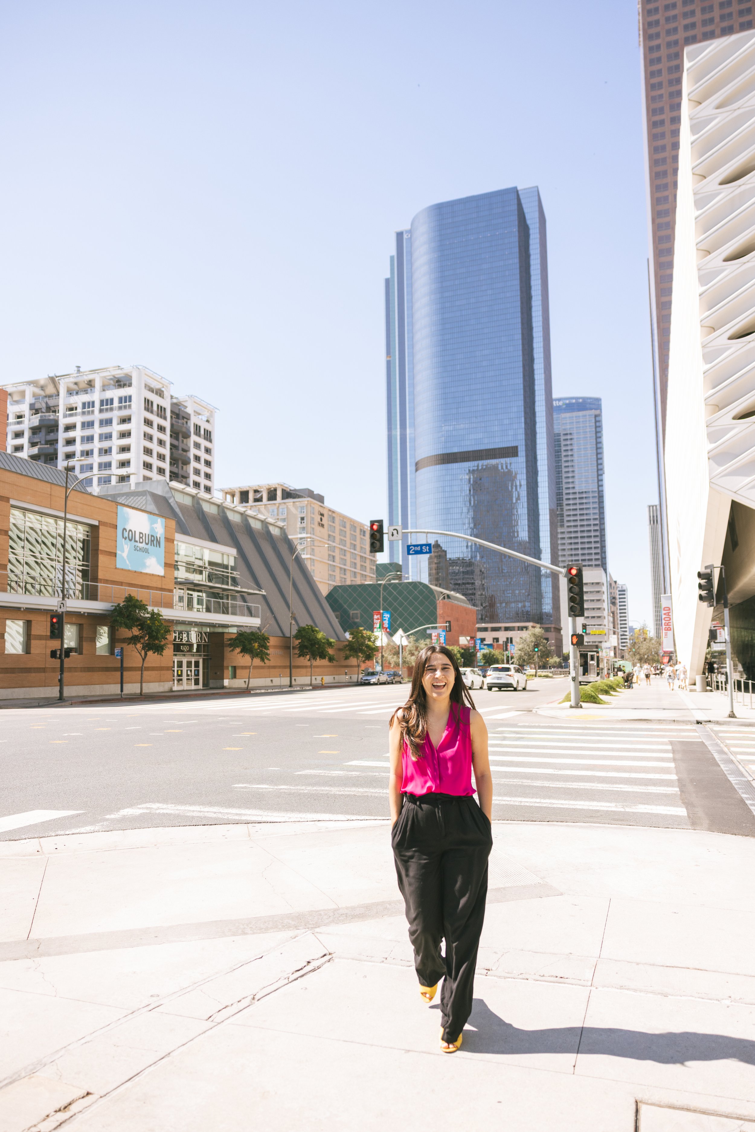A woman in a pink blouse and black pants walking on a city sidewalk with tall buildings and clear blue sky in the background.