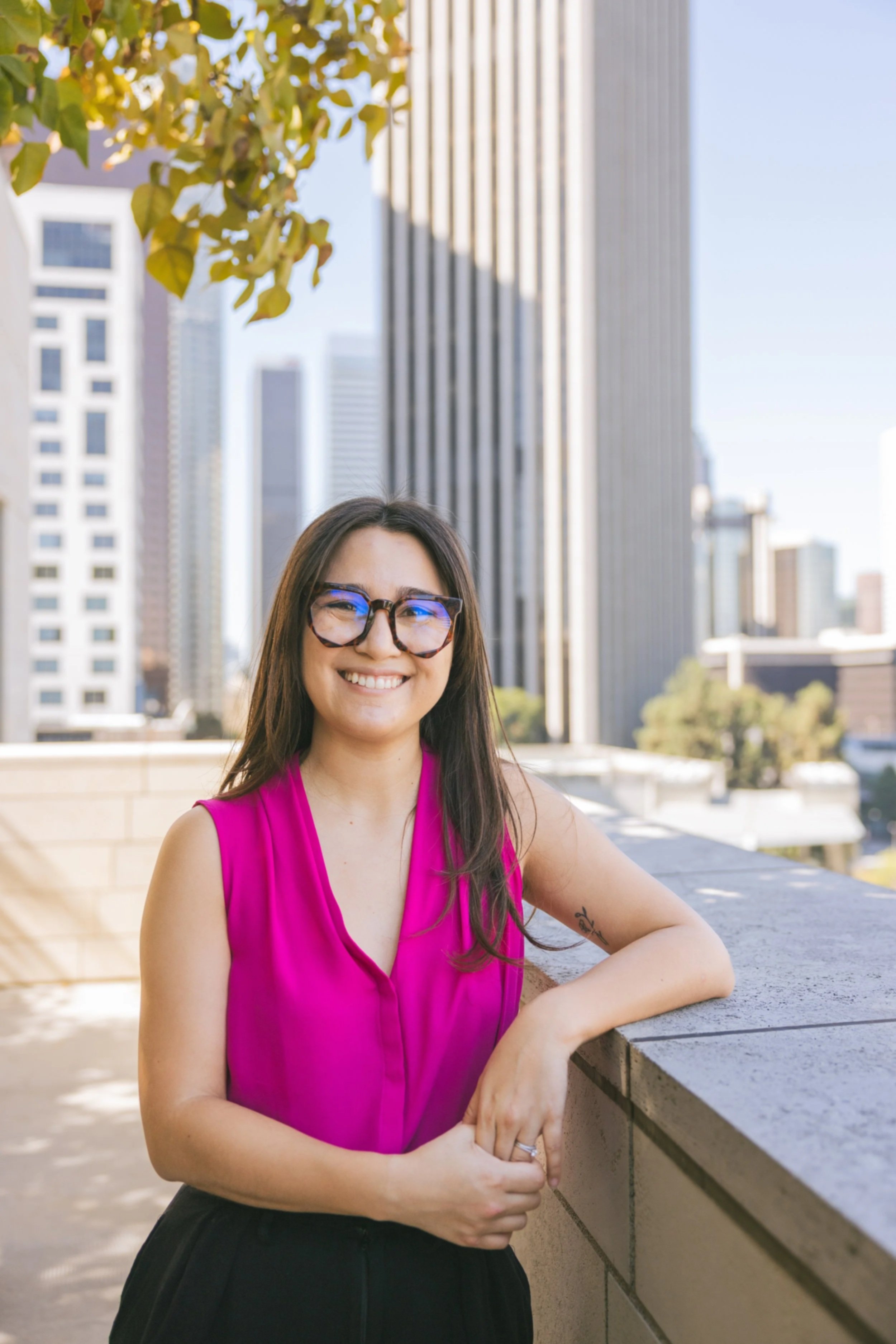 A smiling woman with long dark hair, wearing glasses and a pink sleeveless blouse, leaning on a concrete ledge with an urban cityscape of tall buildings in the background.
